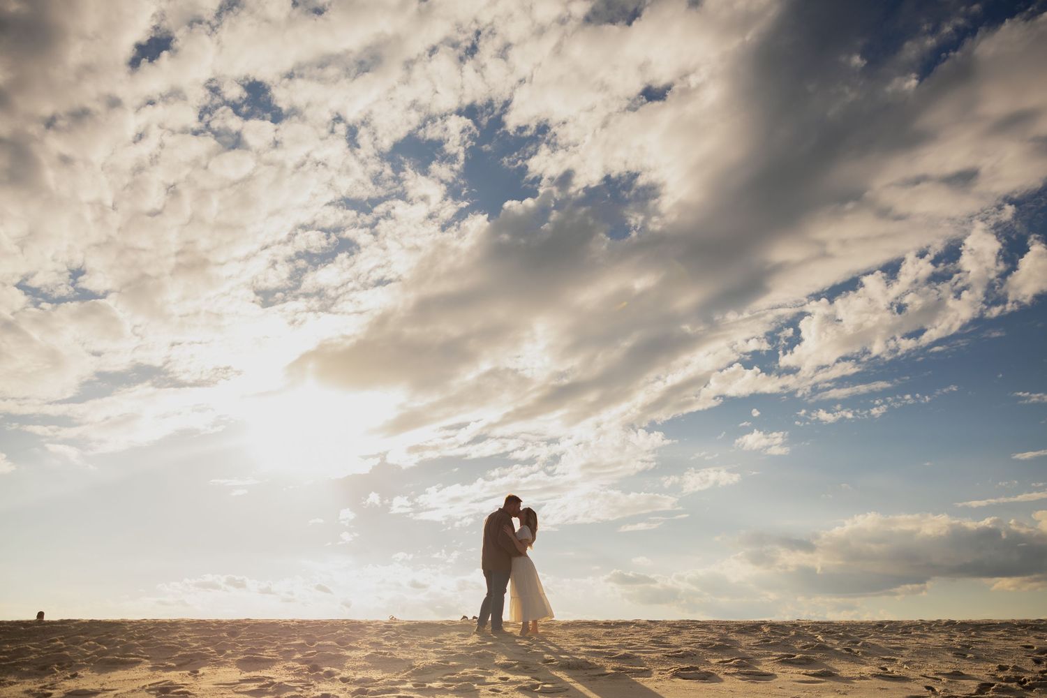 A silhouetted couple embraces on a sandy beach under dramatic clouds during golden hour.