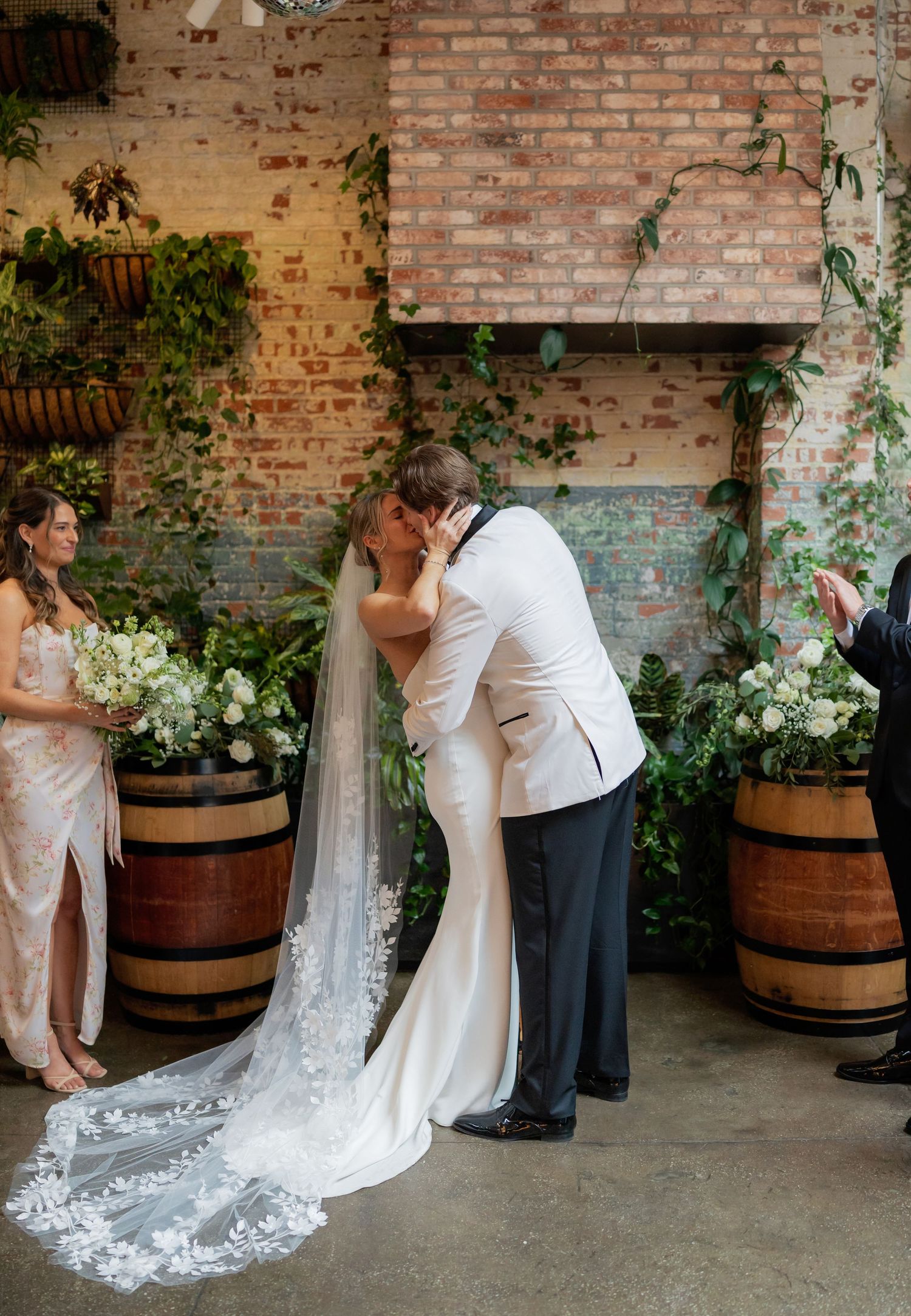 Newlyweds share their first kiss during ceremony in front of exposed brick wall with climbing vines.