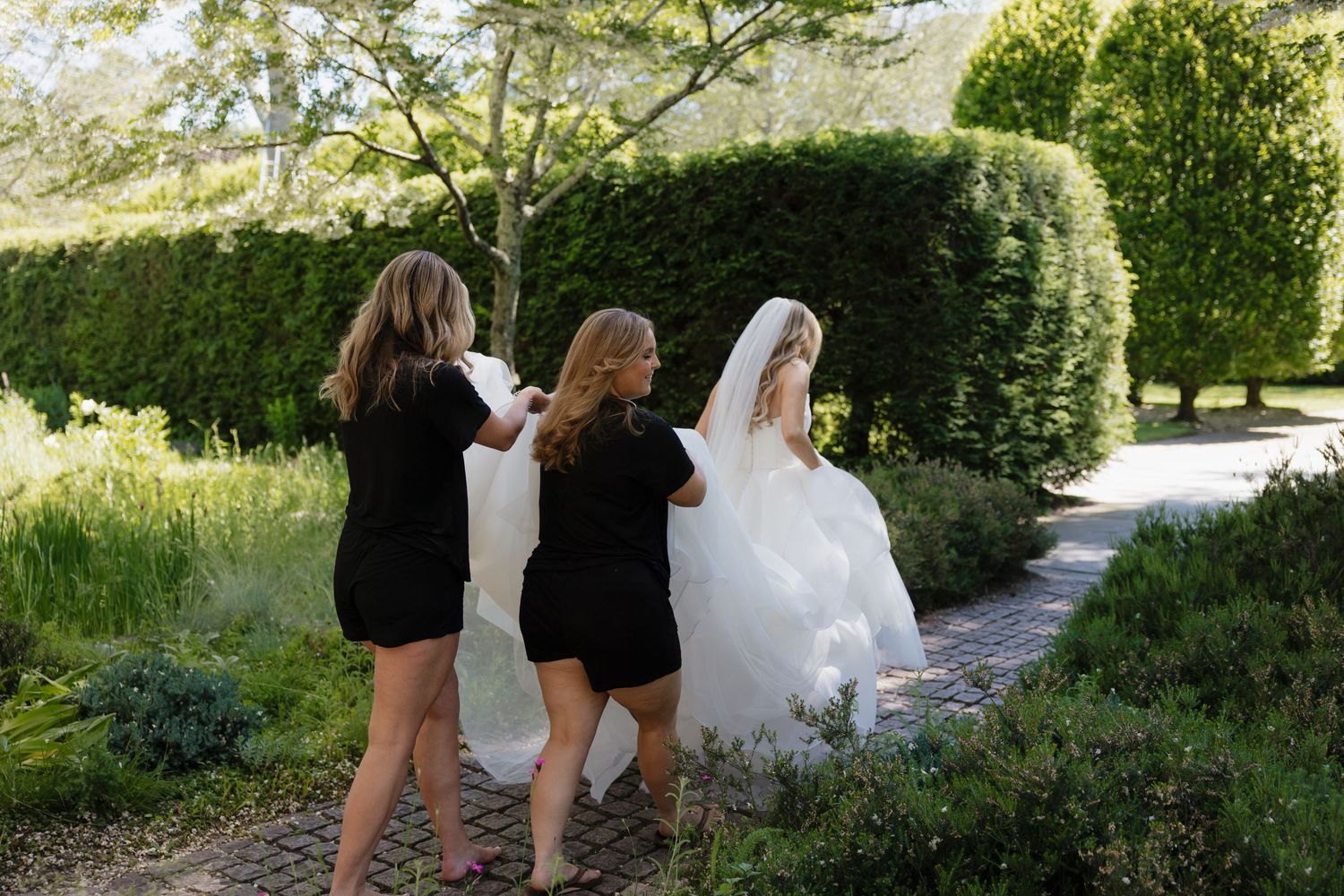 Wedding party walks down a garden path surrounded by lush greenery and trees on a sunny day.