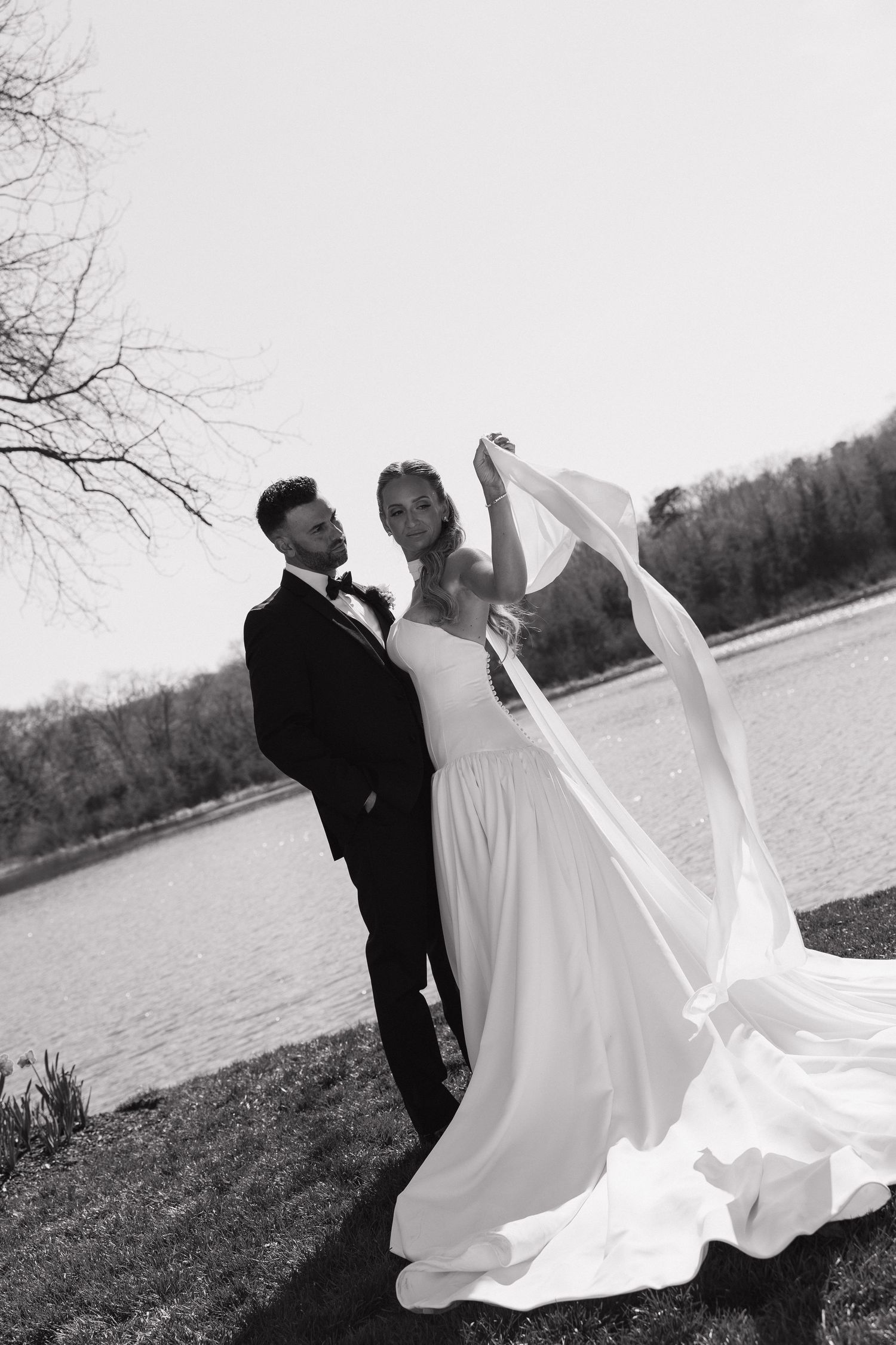 Black and white photo of bride and groom posing by a lake, brides veil flowing in the wind.