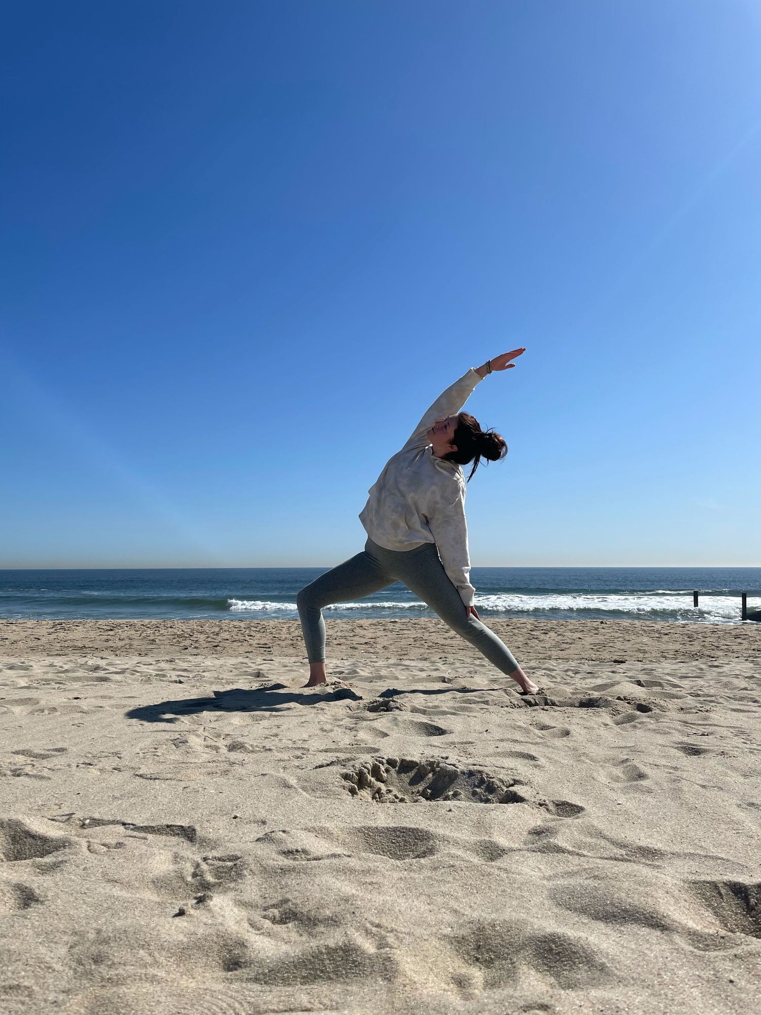 A person in athletic wear performs a yoga pose on a sunny beach with ocean waves in the background.