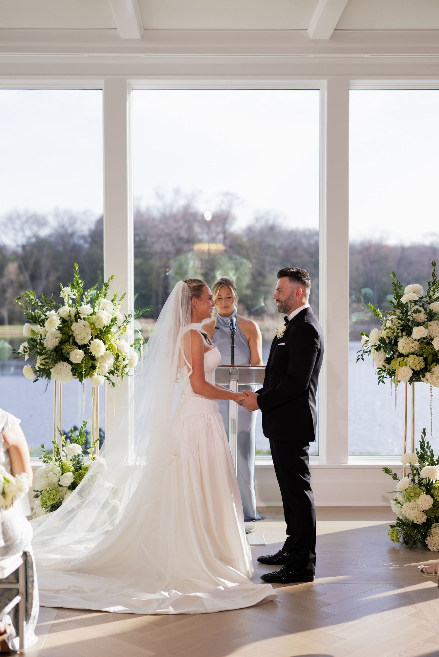 Bride and groom exchange vows at elegant indoor wedding ceremony with white floral arrangements and lake view backdrop.