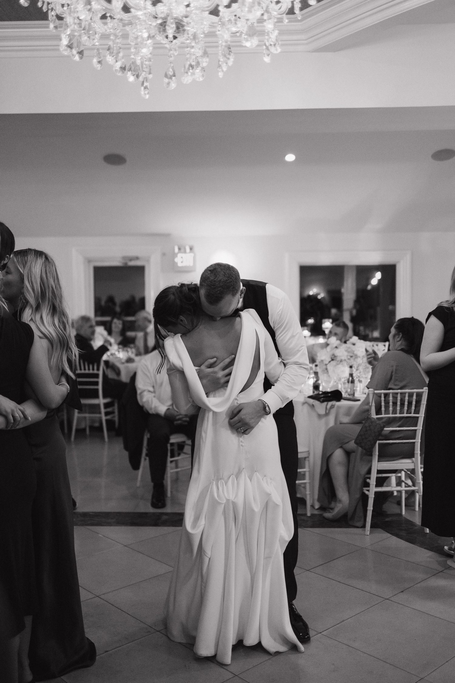 People dance and celebrate at an elegant wedding reception under crystal chandeliers in a white ballroom.