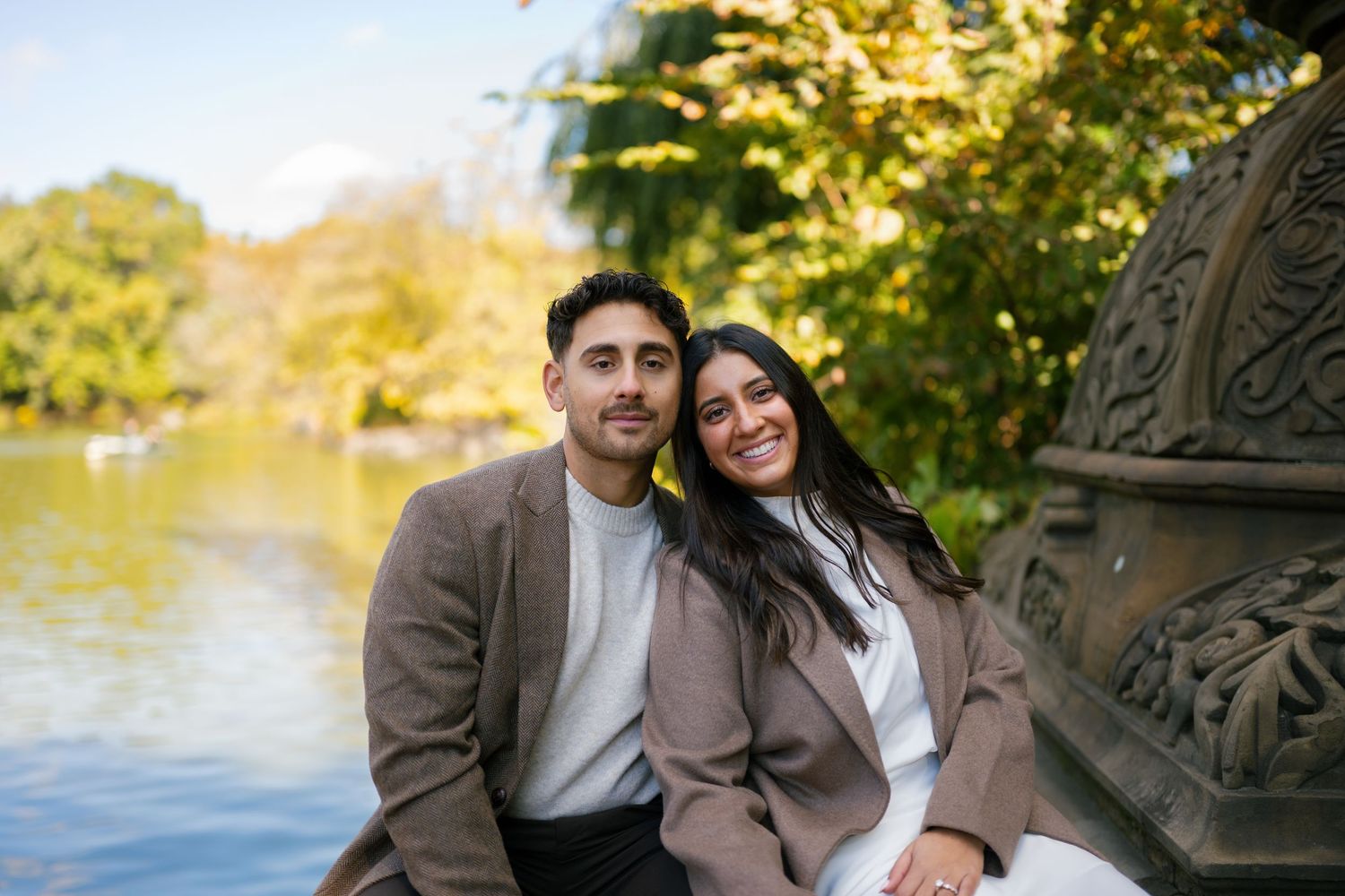 A couple poses together outdoors on a fall day with trees and foliage in the background.