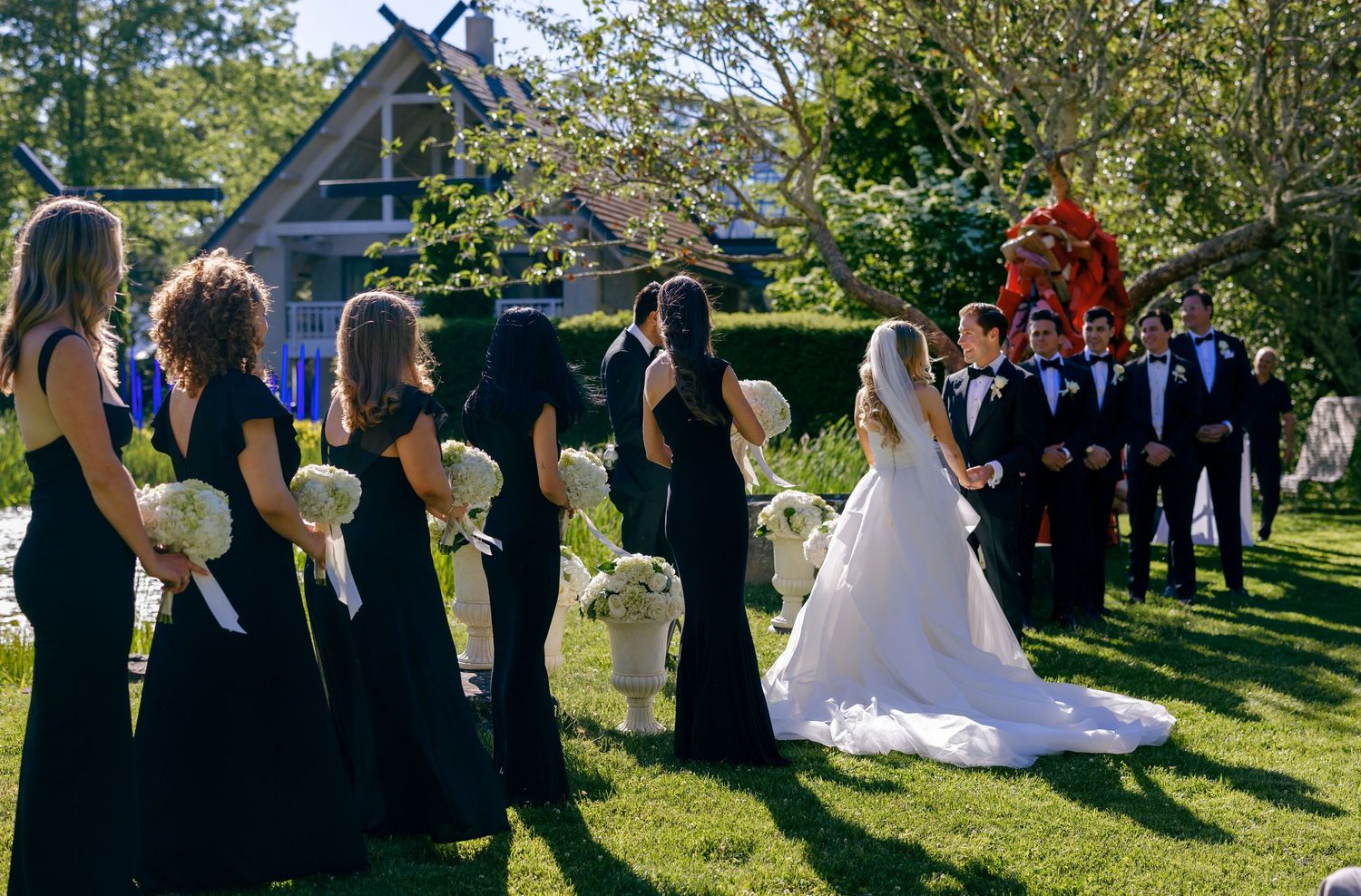 Wedding ceremony guests gather outdoors under trees on a sunny afternoon.
