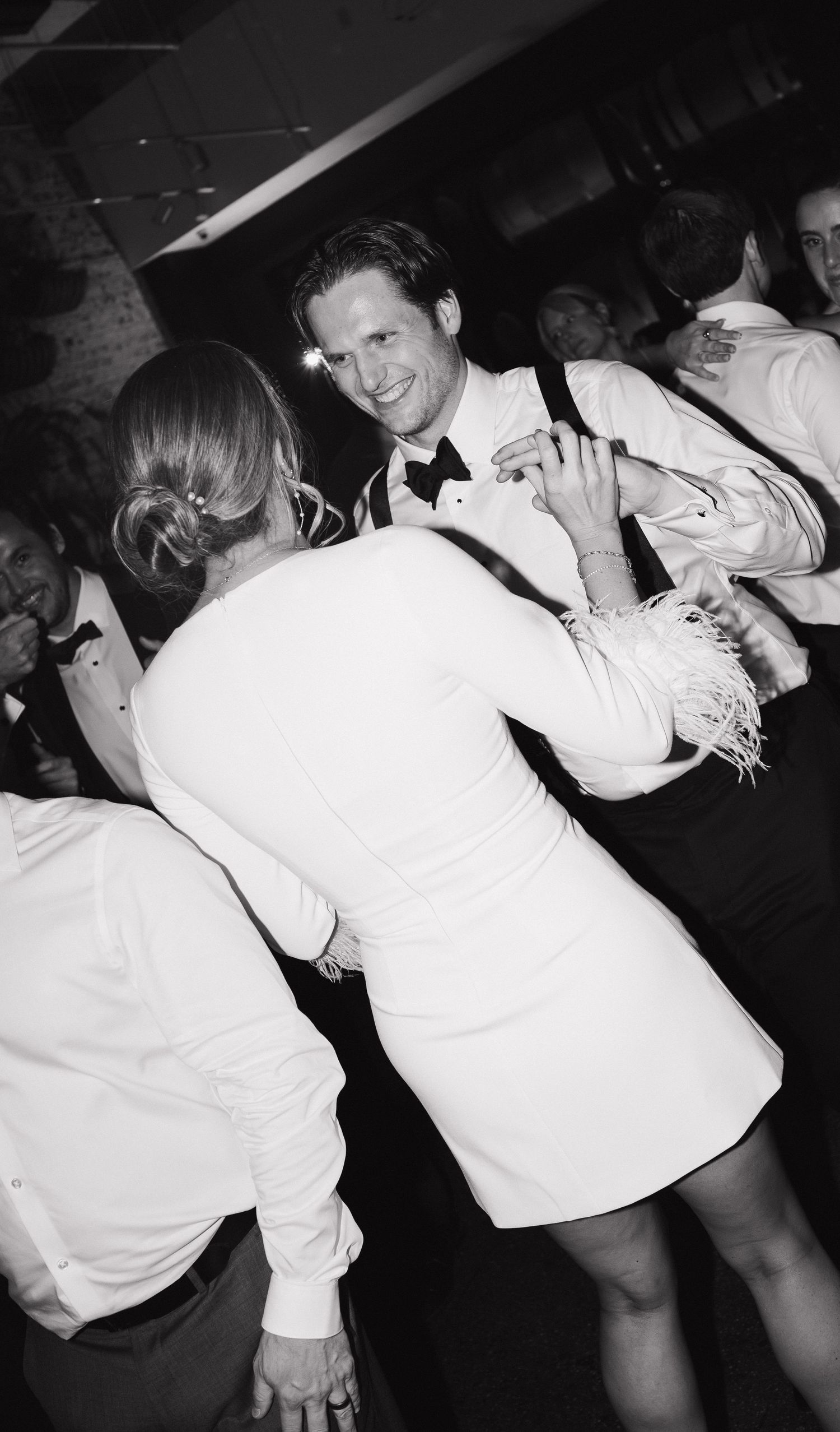 Party guests in white attire dance energetically on a dark dance floor at a wedding reception.