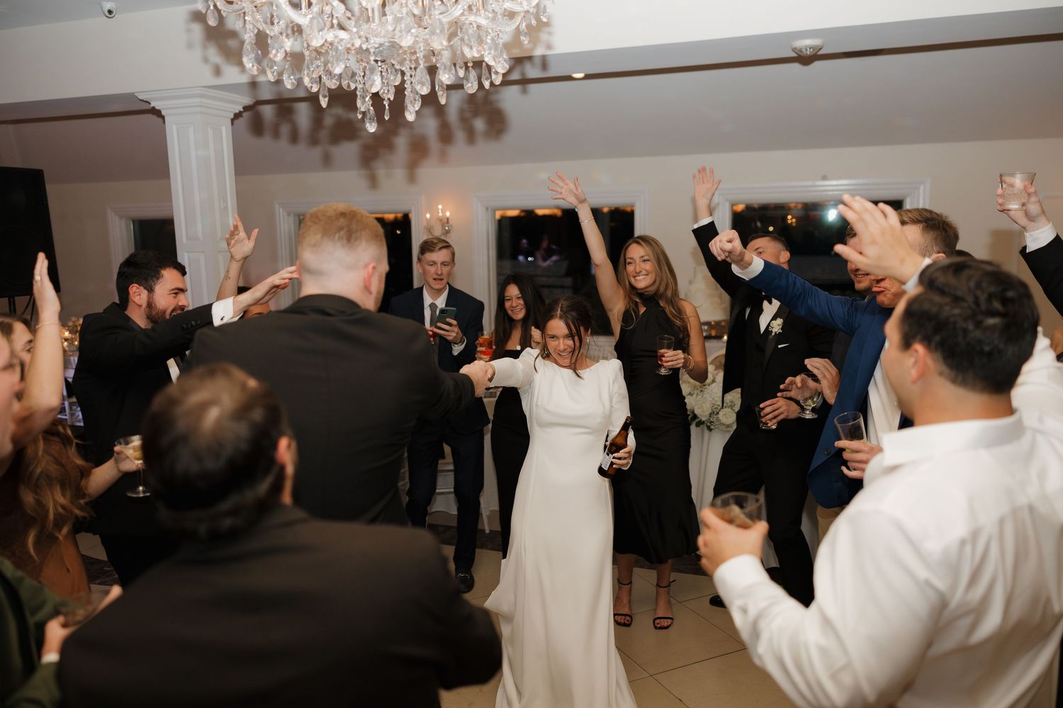 Wedding reception celebration with guests dancing and raising hands under a crystal chandelier.