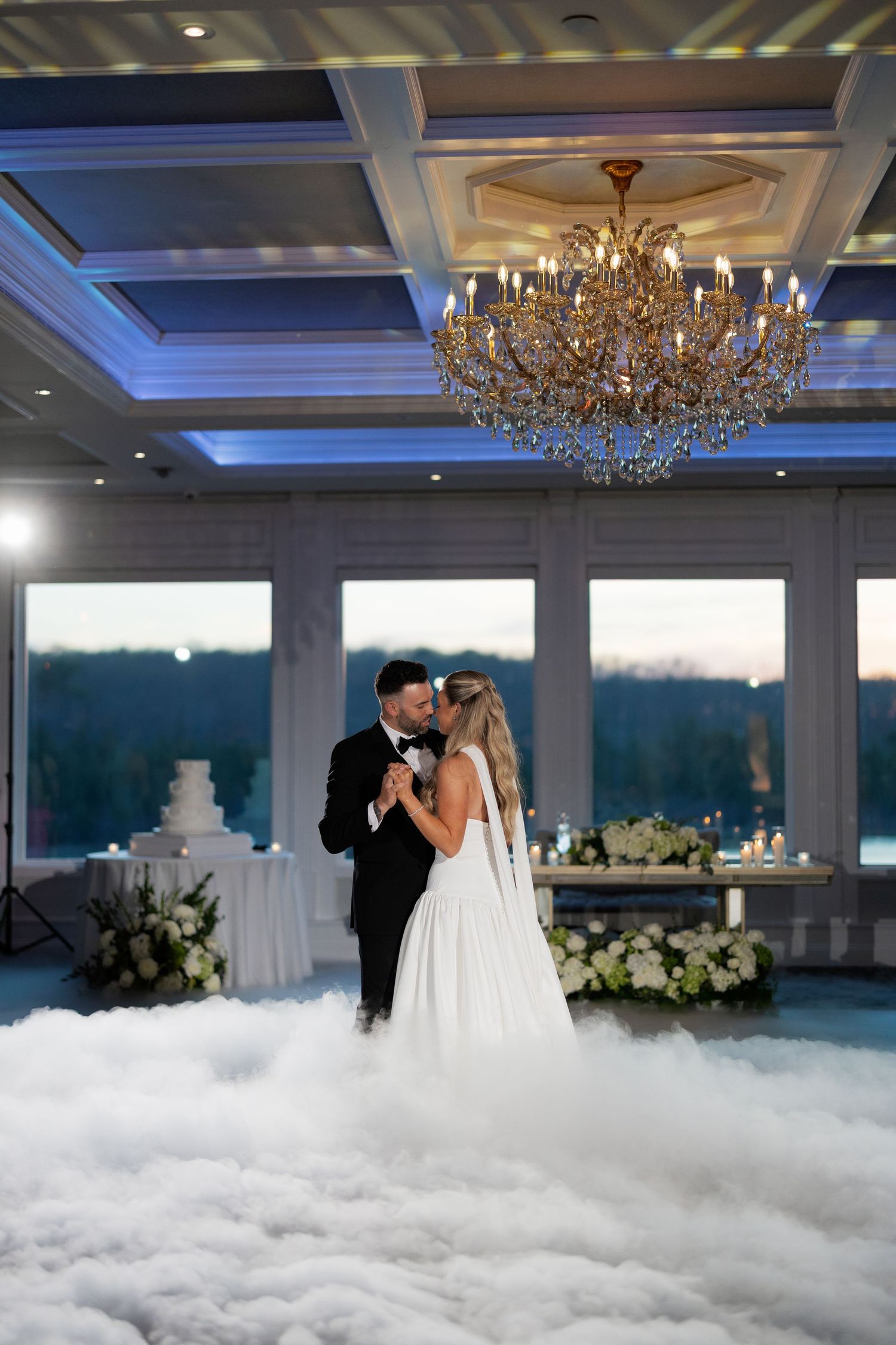 Bride and groom share their first dance surrounded by dramatic fog under a grand chandelier at their wedding reception.