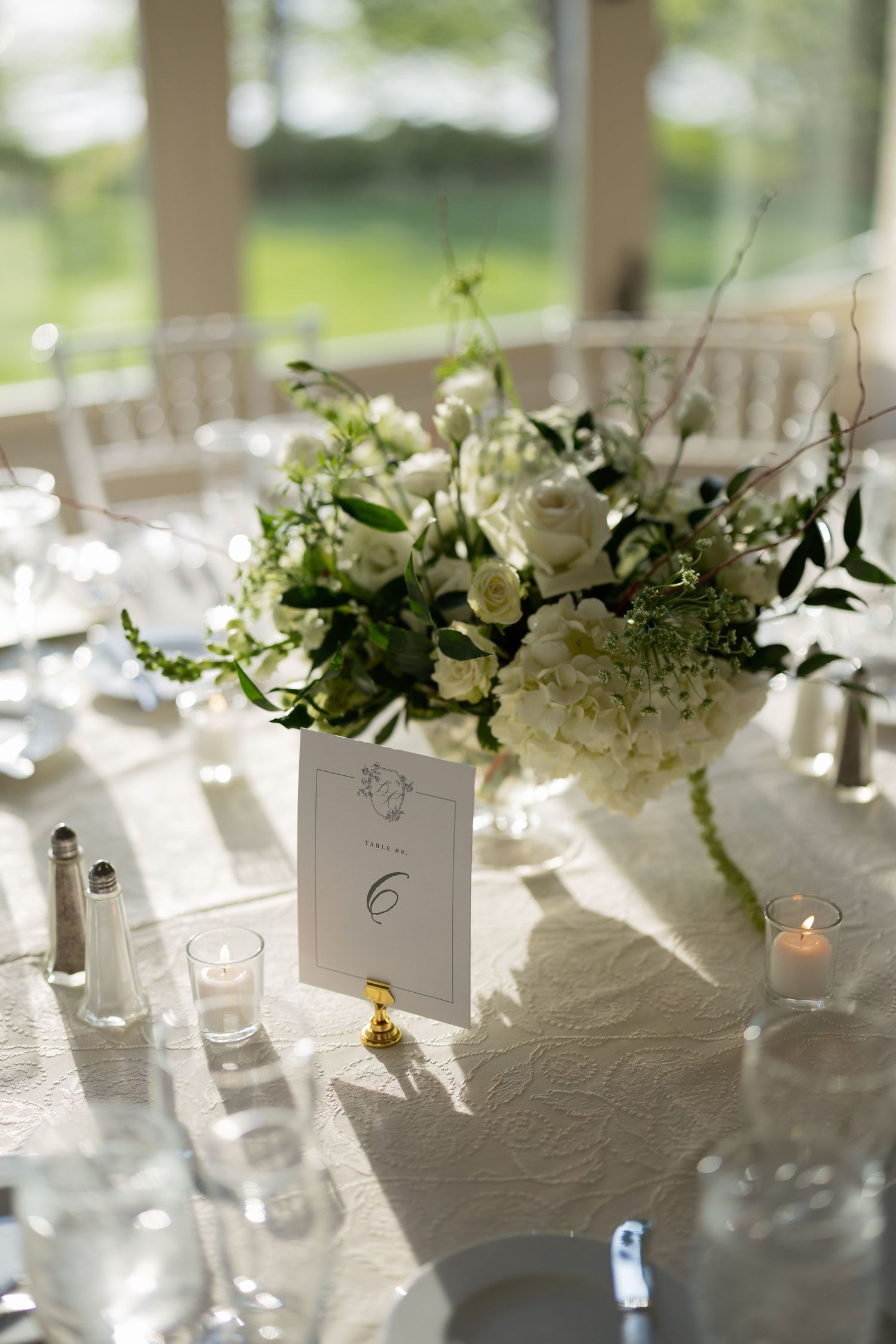 Elegant white floral centerpiece and table number arrangement at a wedding reception with natural lighting.