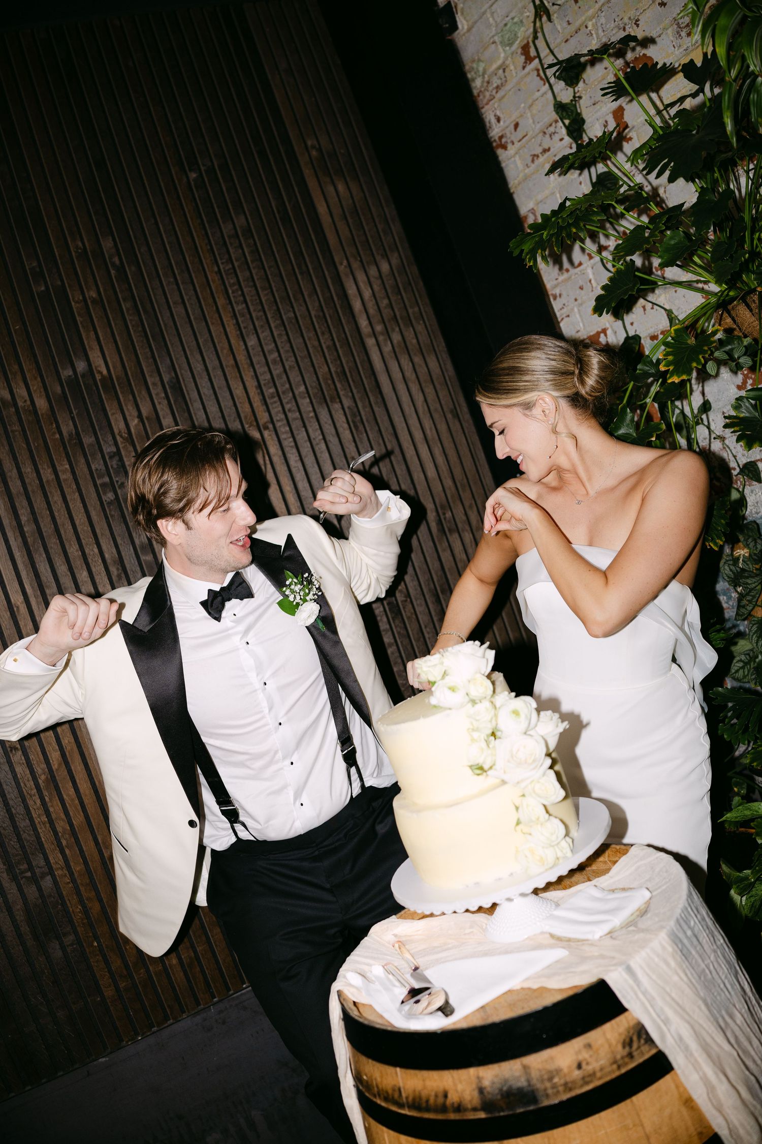 Wedding couple playfully poses while cutting their elegant white wedding cake placed on a wooden barrel.
