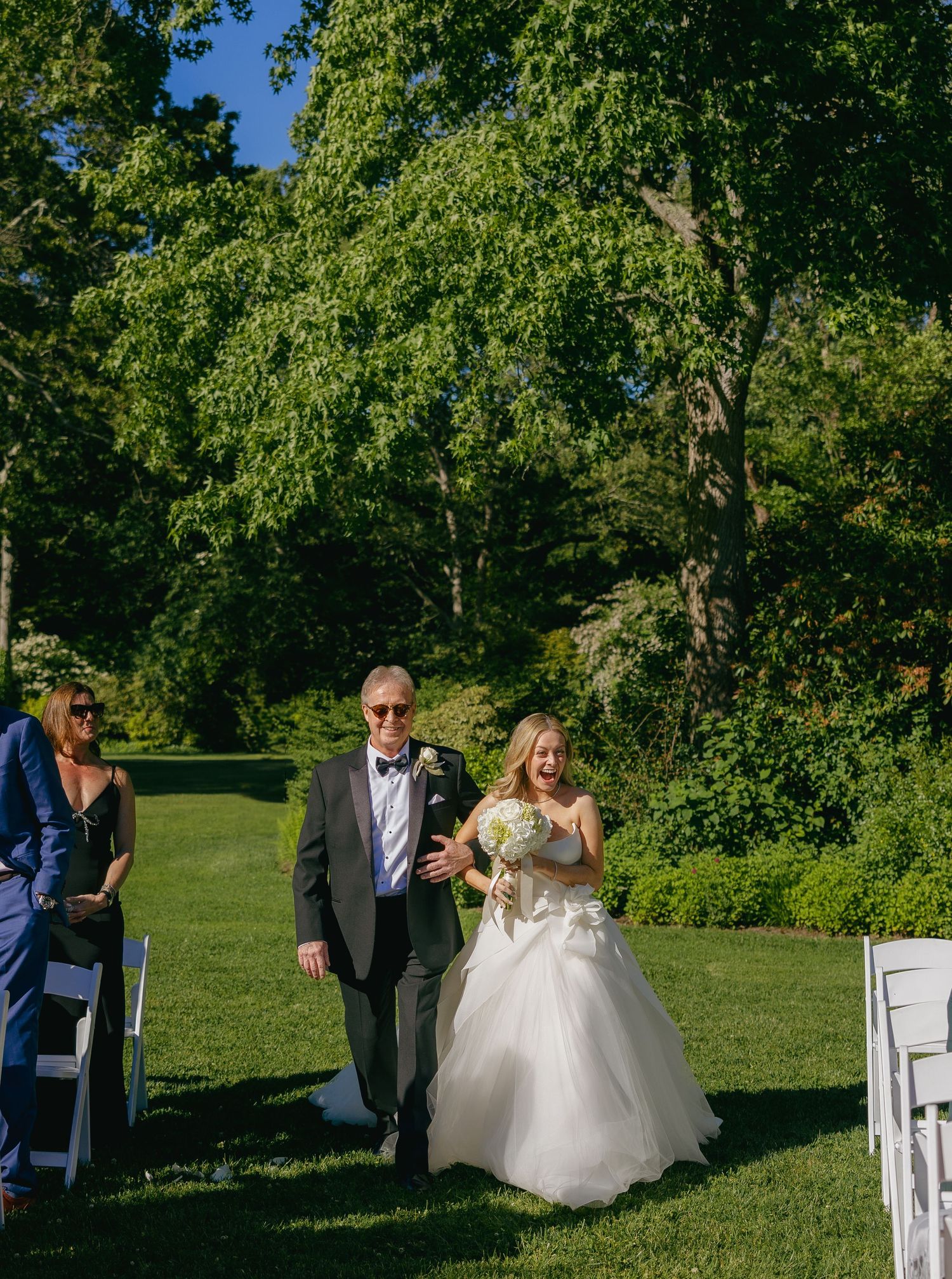 Wedding ceremony setup outdoors with white chairs and natural greenery backdrop.