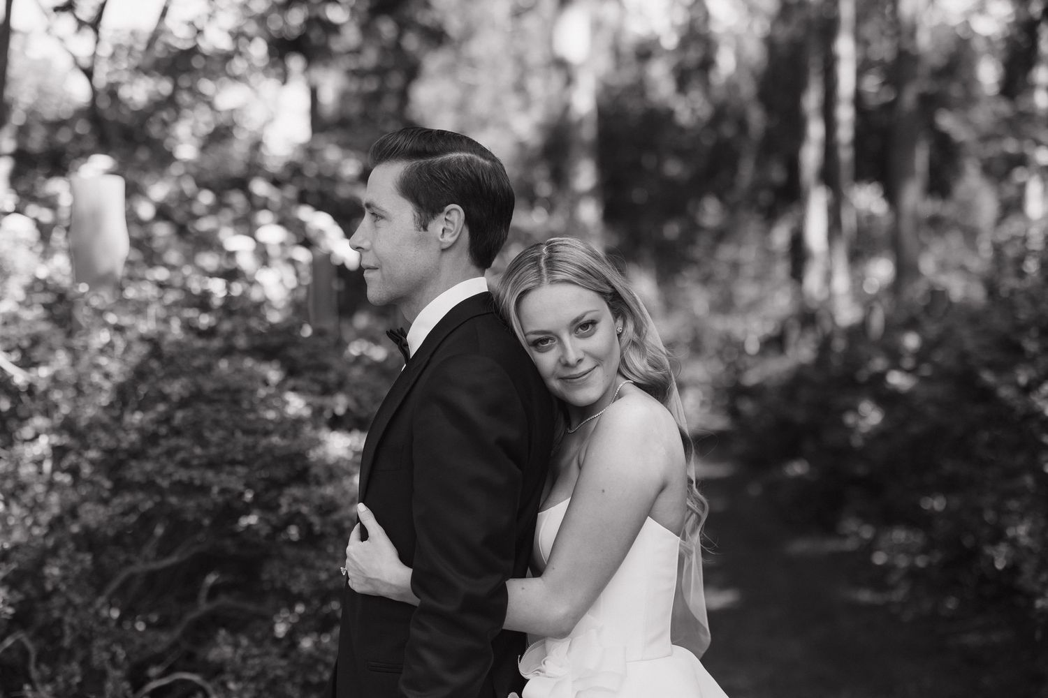 A romantic black and white wedding portrait shows a couple embracing in a wooded setting.