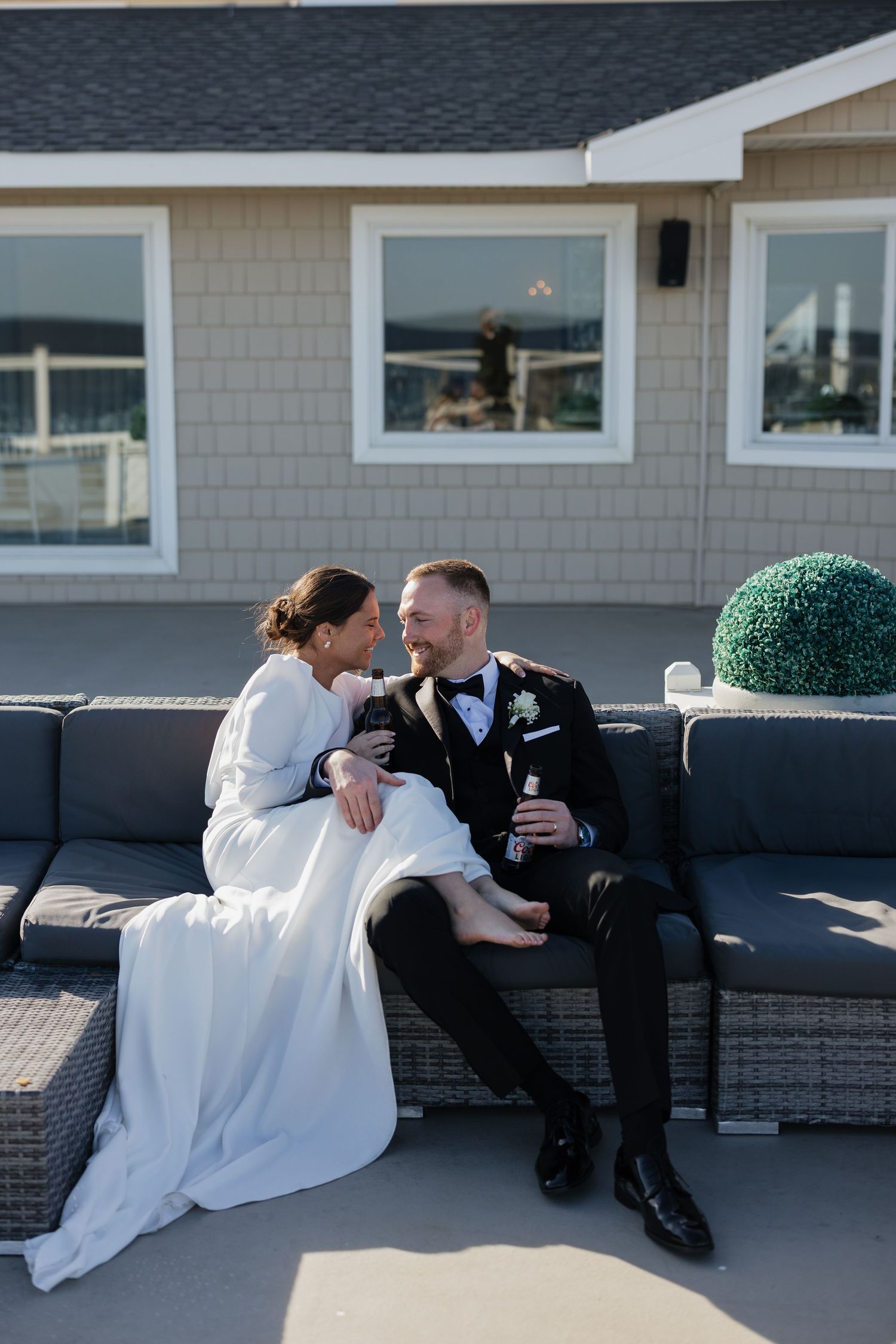 A couple in wedding attire share intimate moments while sitting on a bench outside a coastal building.