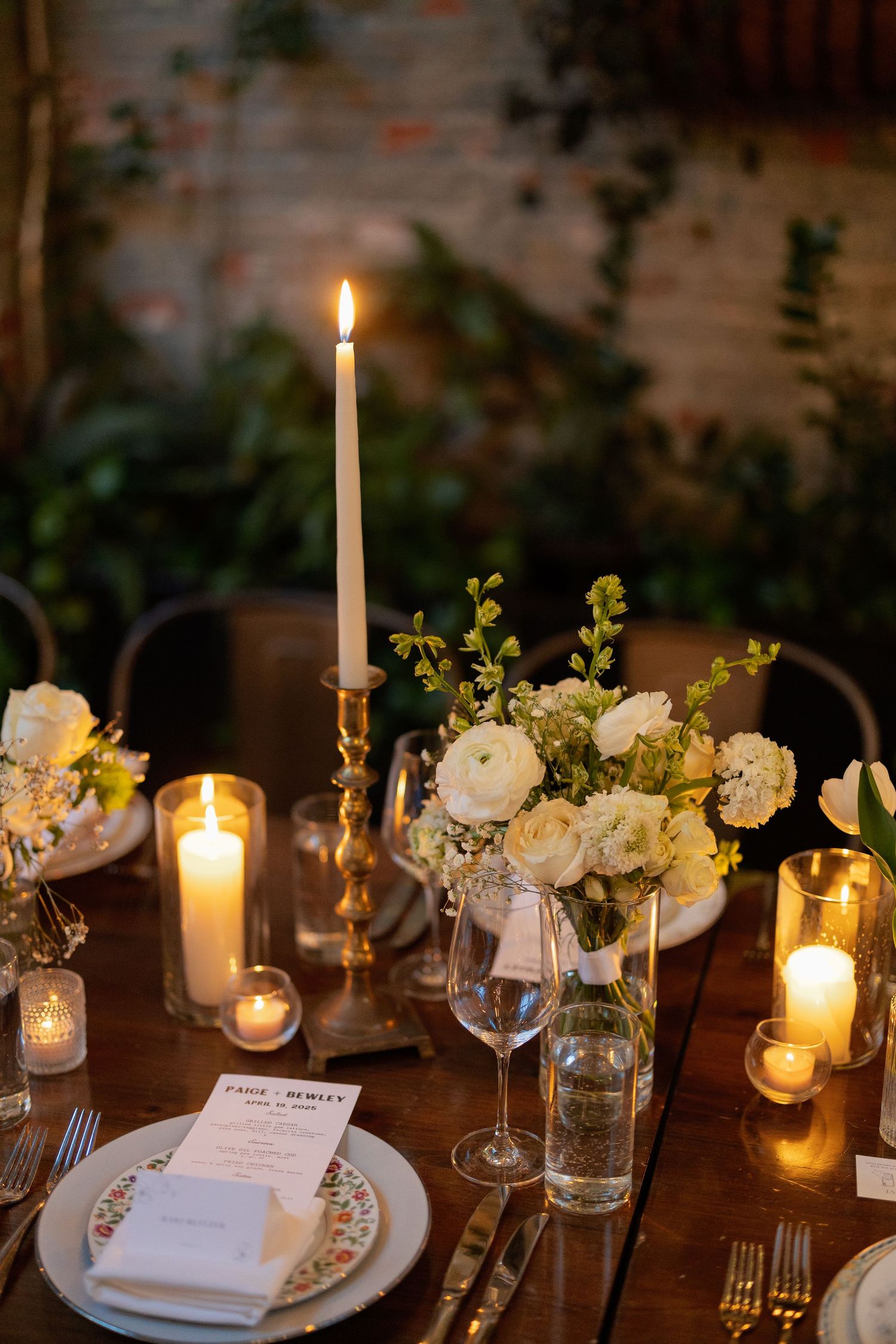Elegant wedding table setting with white flowers, glowing candles, and crystal glassware on dark wooden tables.
