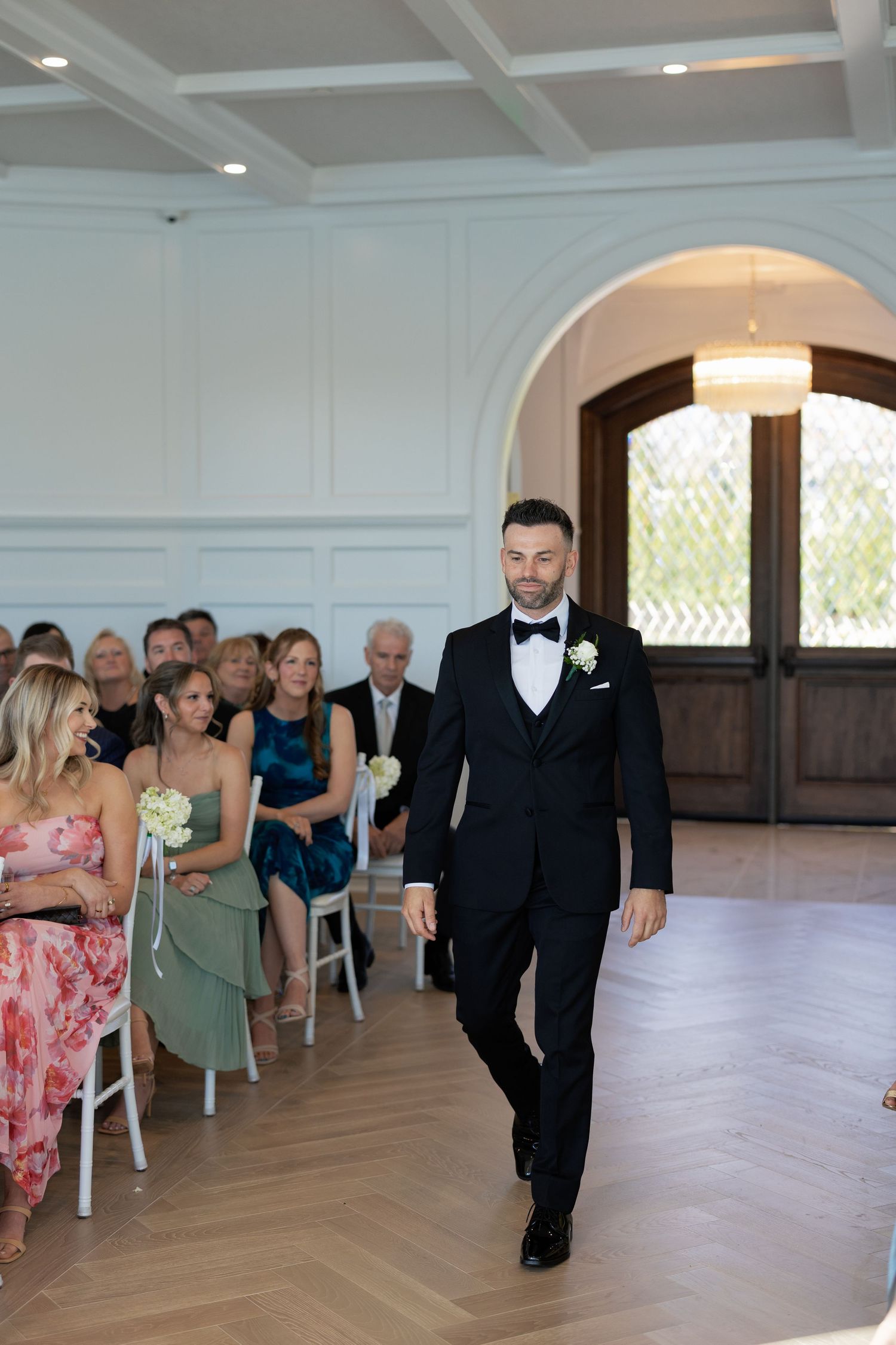 Dapper groom in black tuxedo walking down the aisle at an elegant indoor wedding ceremony.
