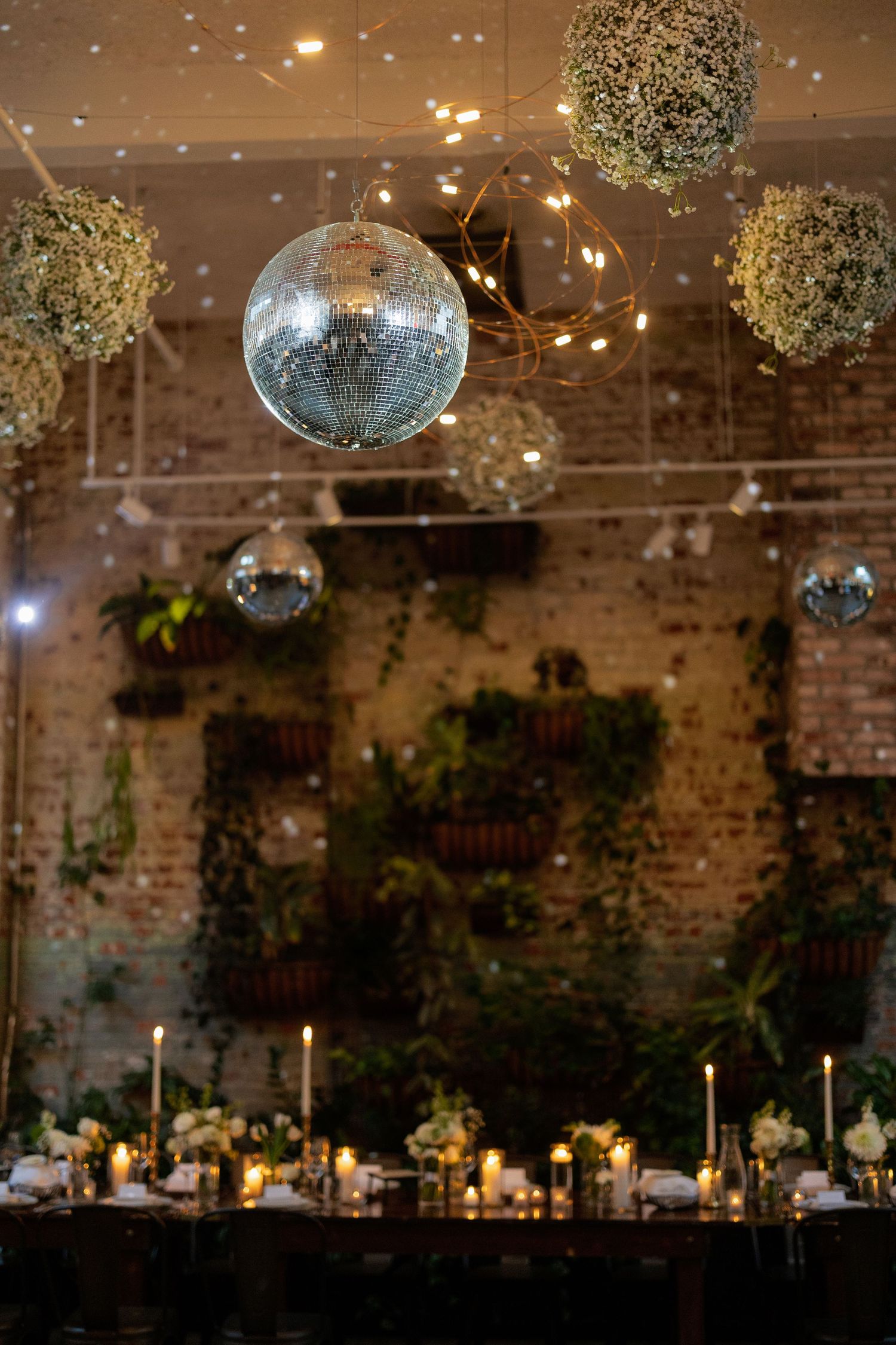 Disco balls and floral arrangements hang from ceiling above candlelit dining tables in an industrial brick venue.