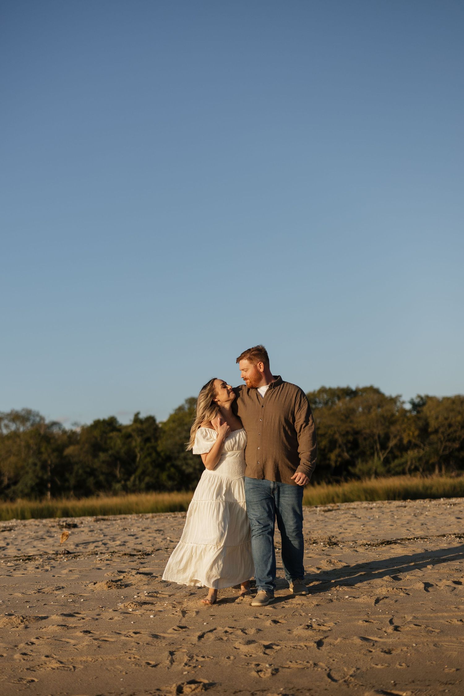 A couple stands together on a sandy beach at golden hour with trees in the background.