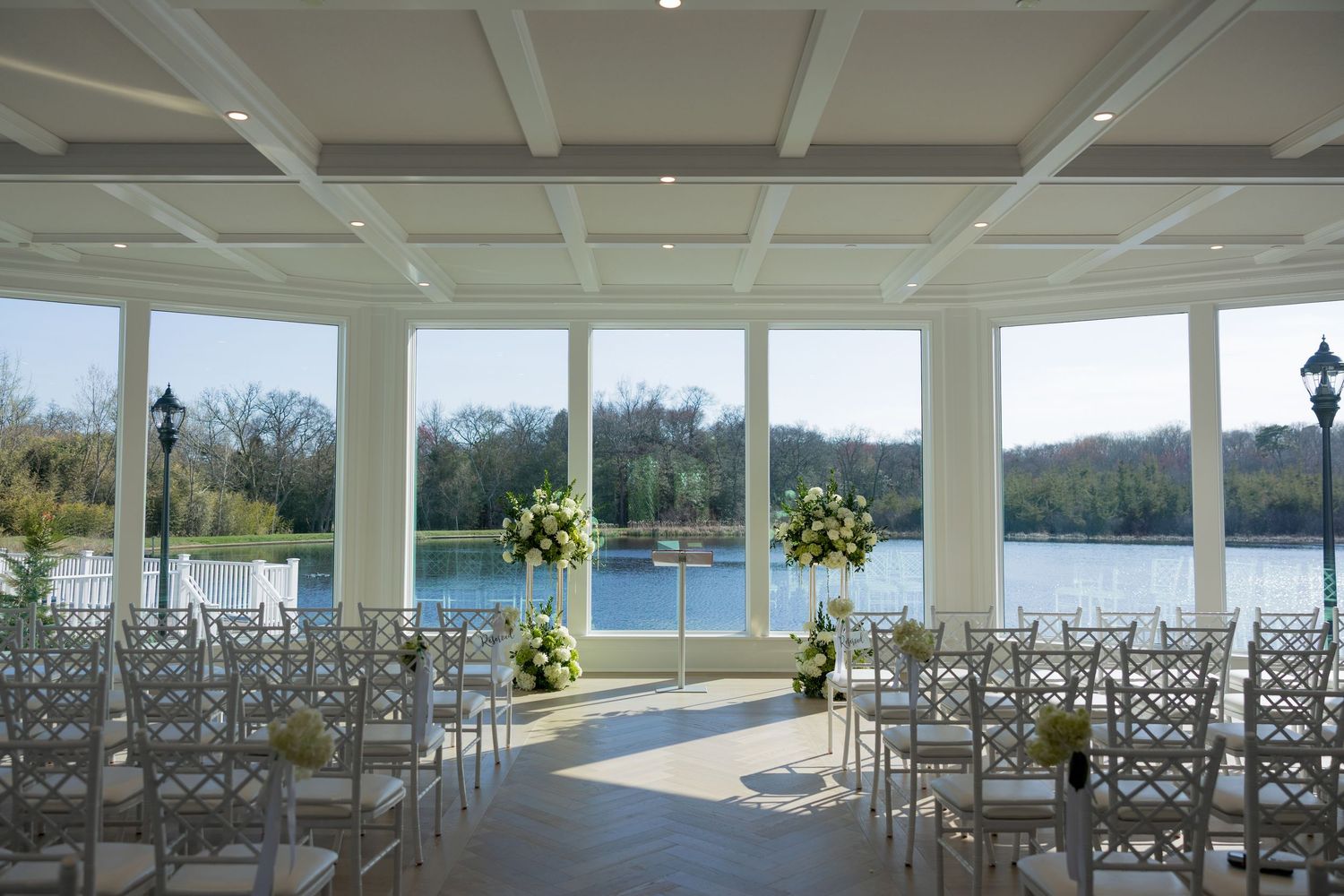 Elegant wedding venue with white chairs, floral arrangements, and panoramic windows overlooking a serene lake.