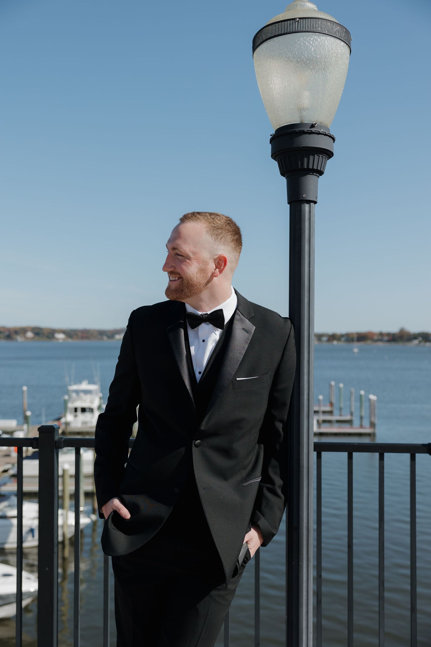 A person in a black tuxedo stands by a waterfront dock next to a vintage lamp post on a sunny day.