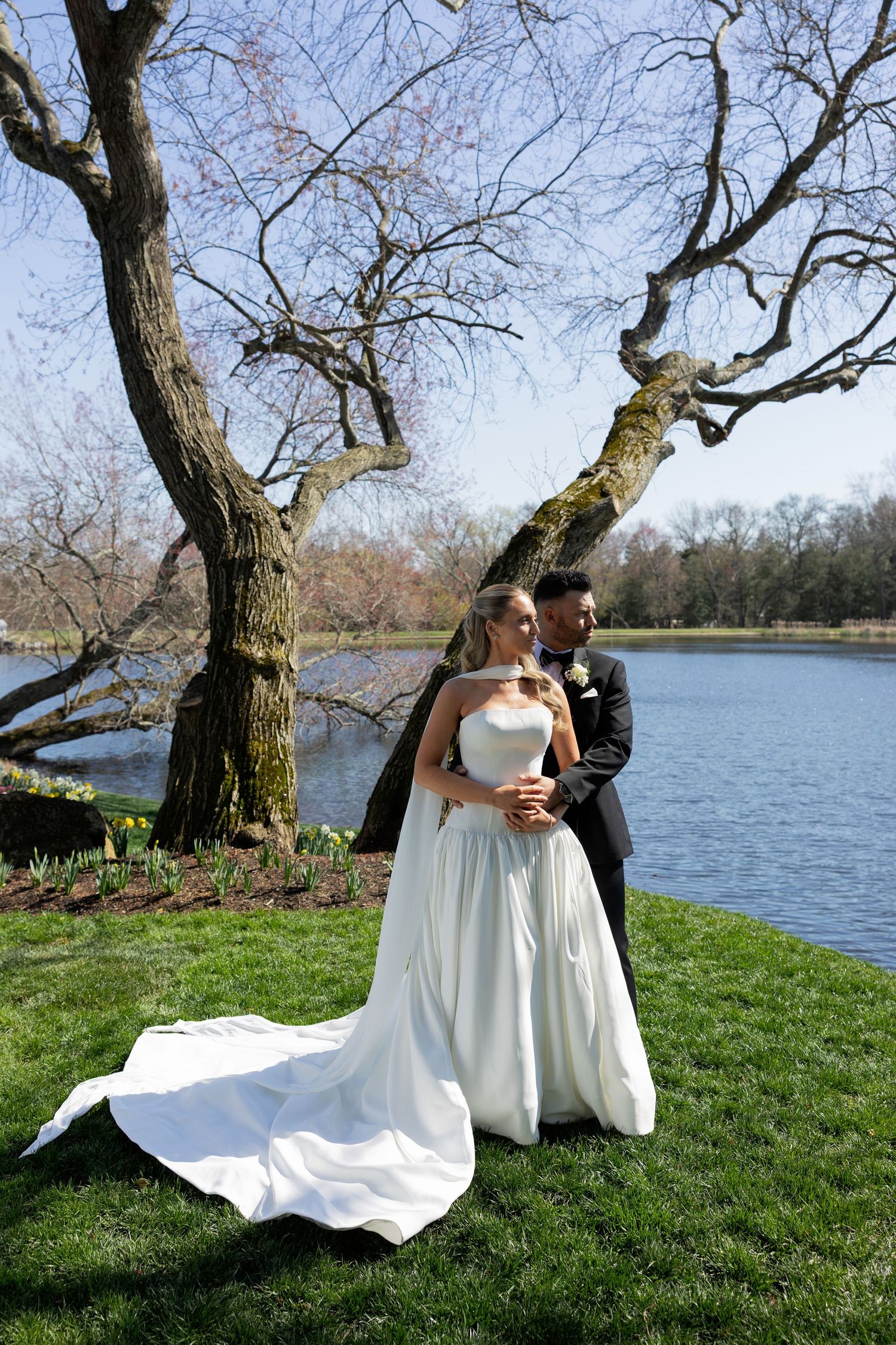 Bride and groom posing romantically by a serene lake under bare trees on their wedding day.