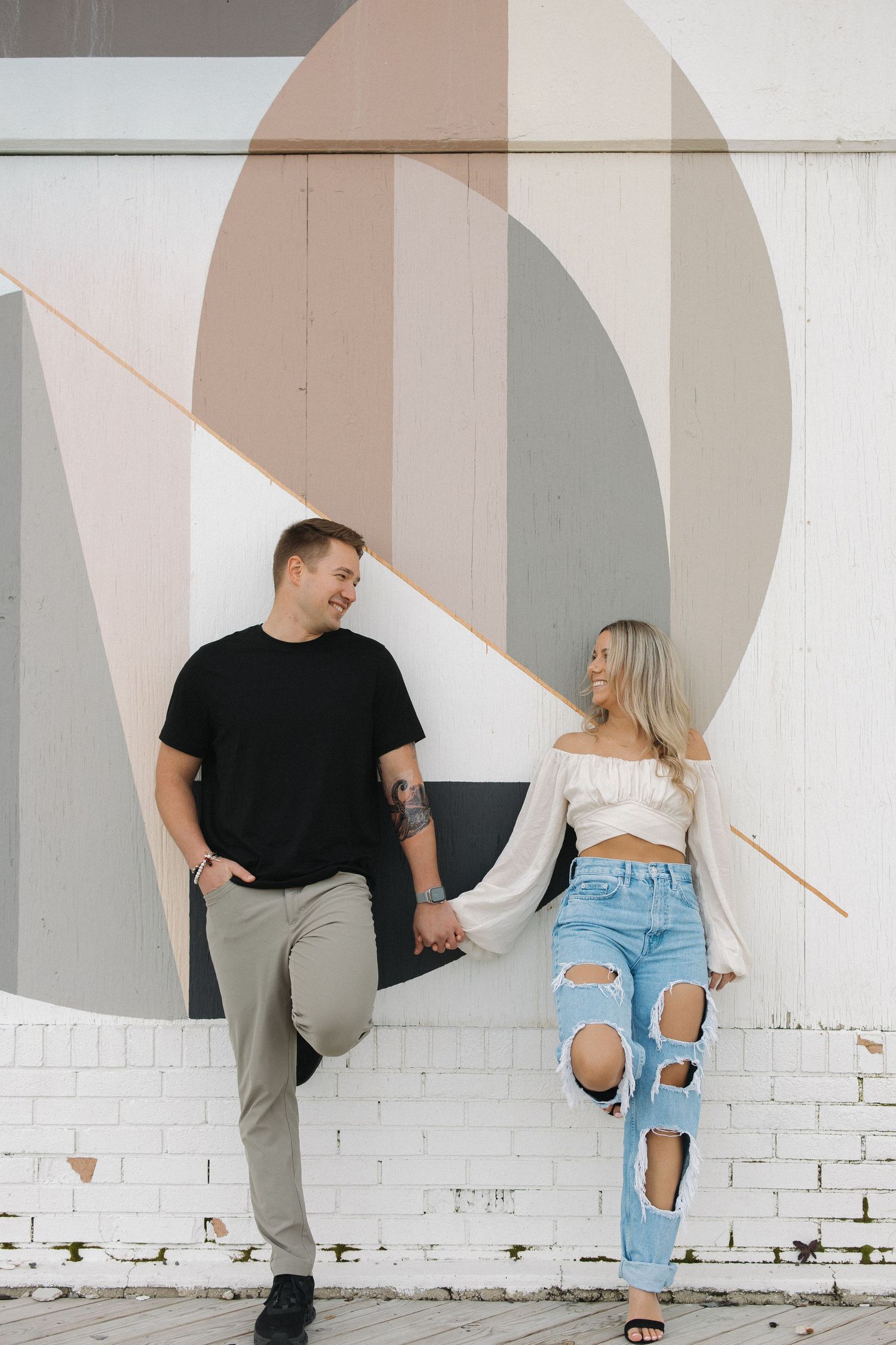Modern couple posing against white brick wall with abstract geometric painted shapes in stylish casual outfits.