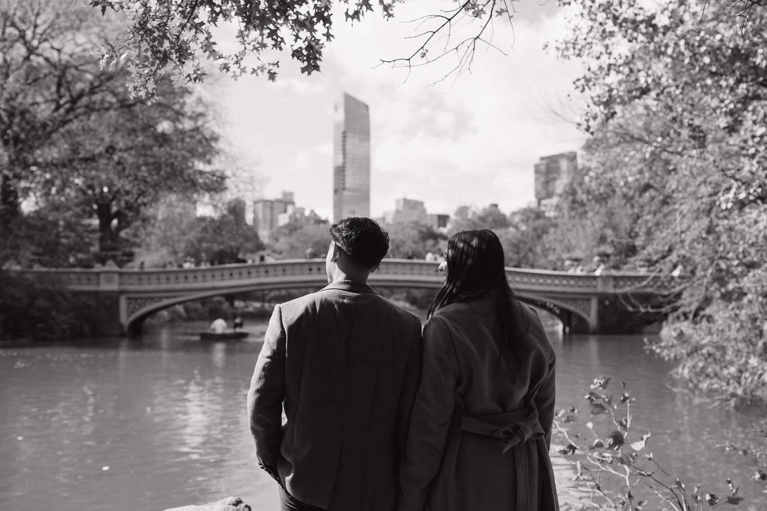 Two people sit together on a bench overlooking Central Park lake and Bow Bridge on an autumn afternoon in New York City.