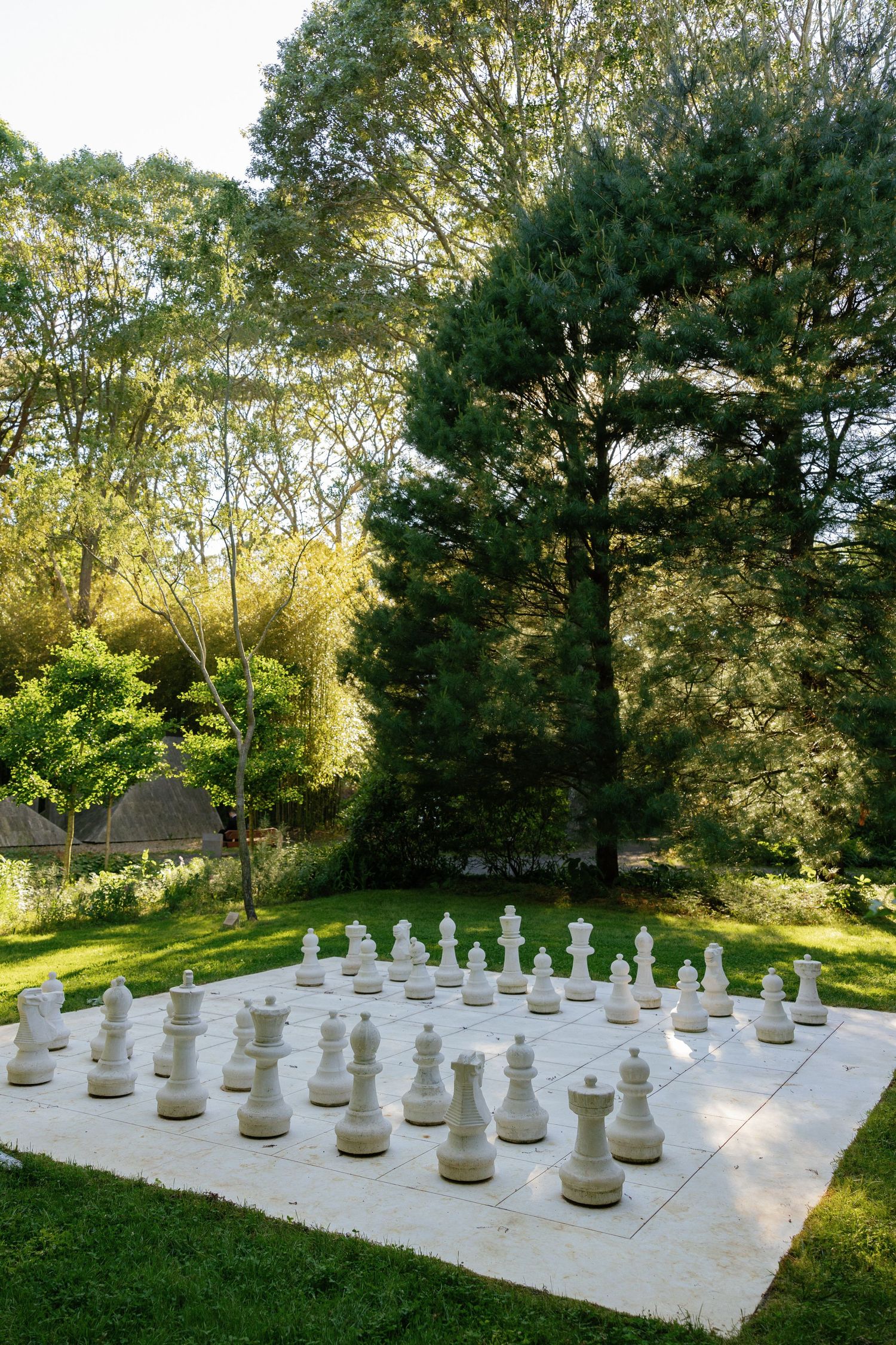 Large outdoor chess set with white pieces arranged on a concrete board surrounded by lush garden landscaping.