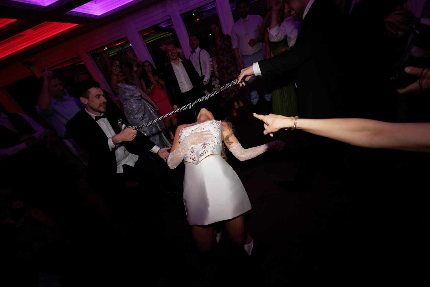 Bride in short white lace dress doing the limbo under a stick at vibrant wedding reception party.