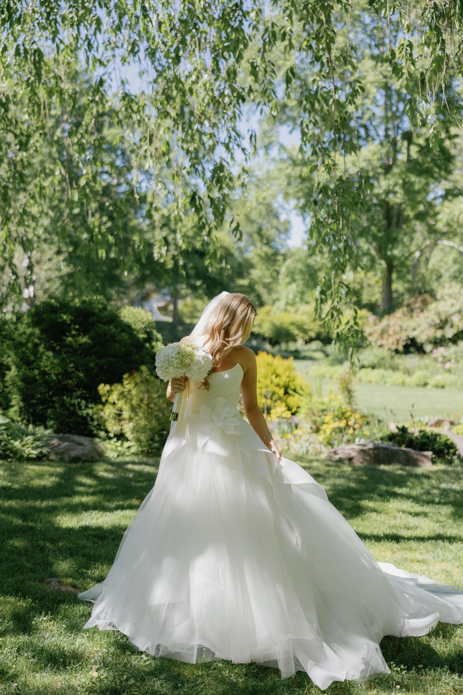 A flowing white wedding dress billows in the breeze under weeping willow trees in a garden setting.