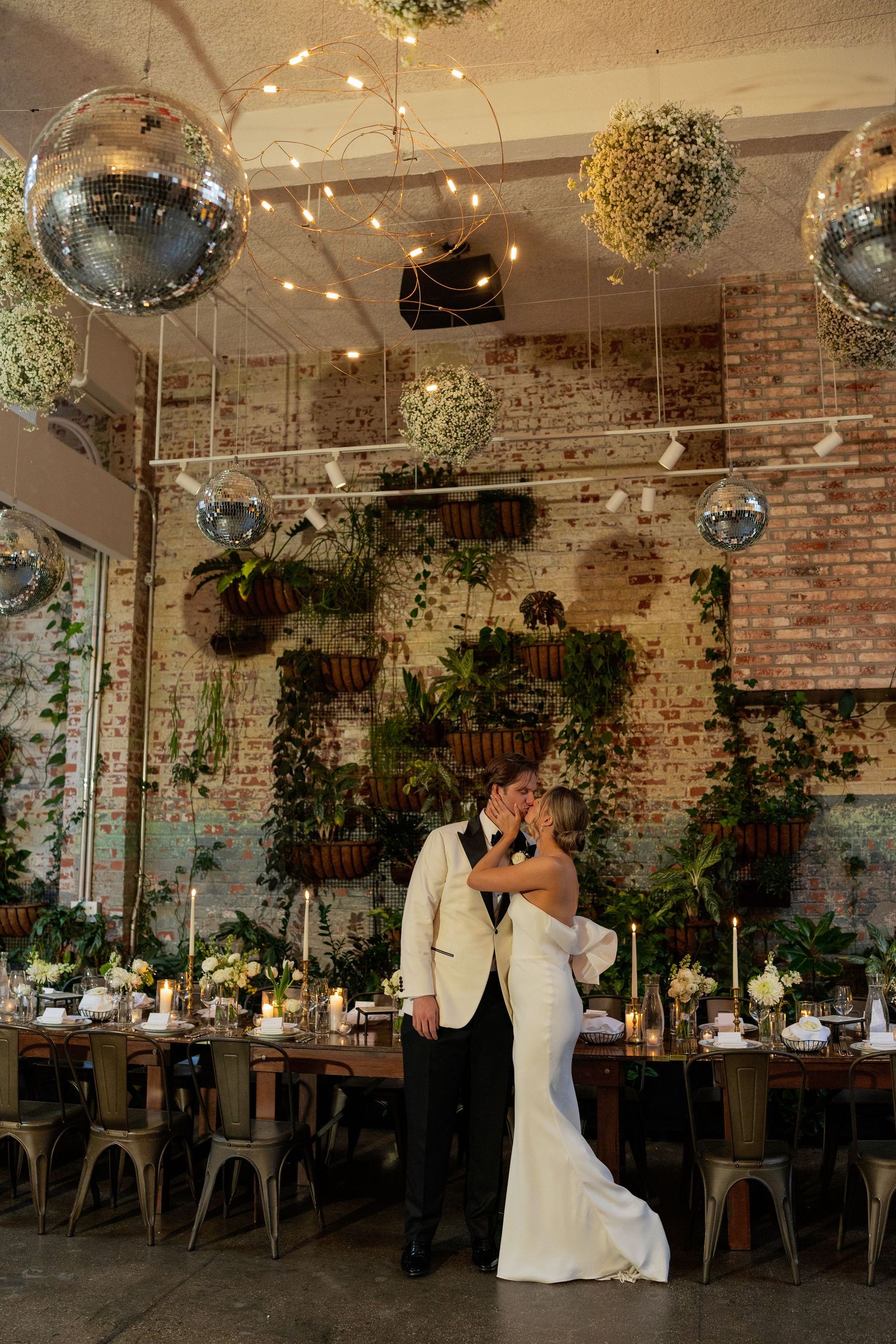 Wedding reception venue features exposed brick walls, hanging disco balls, and lush greenery decorations.