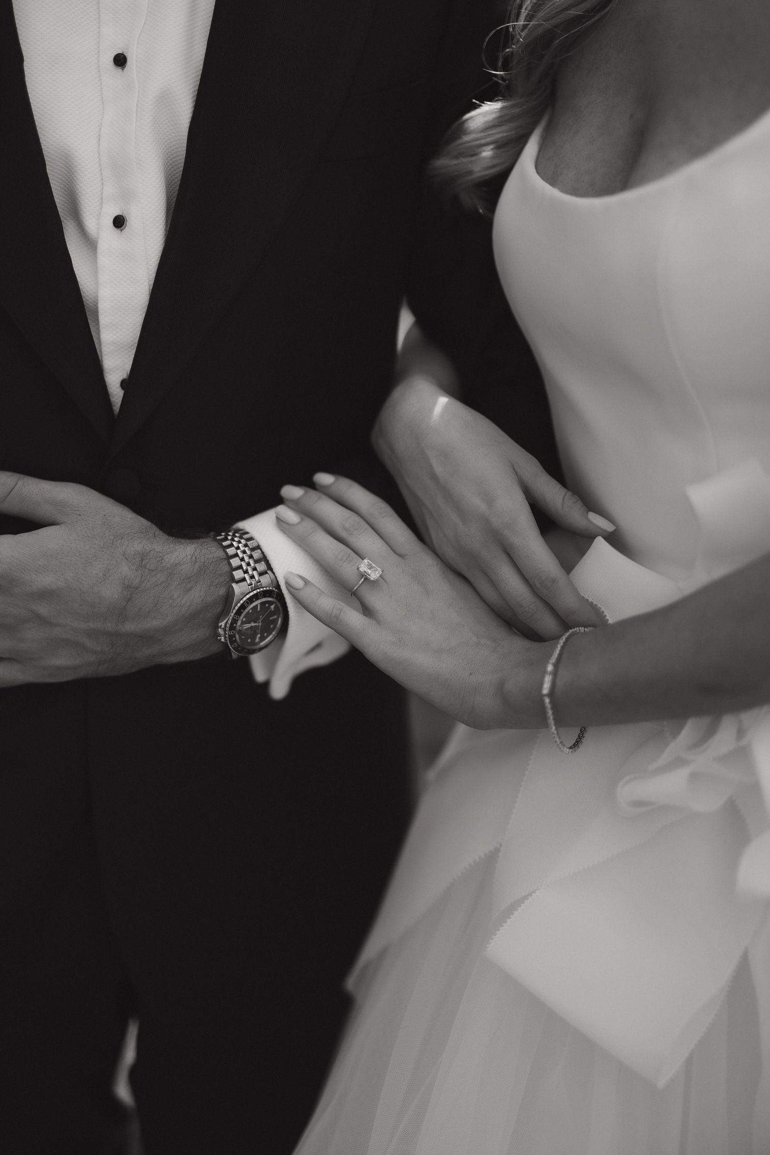 Black and white close-up of newlyweds' hands showing wedding rings and formal attire details.
