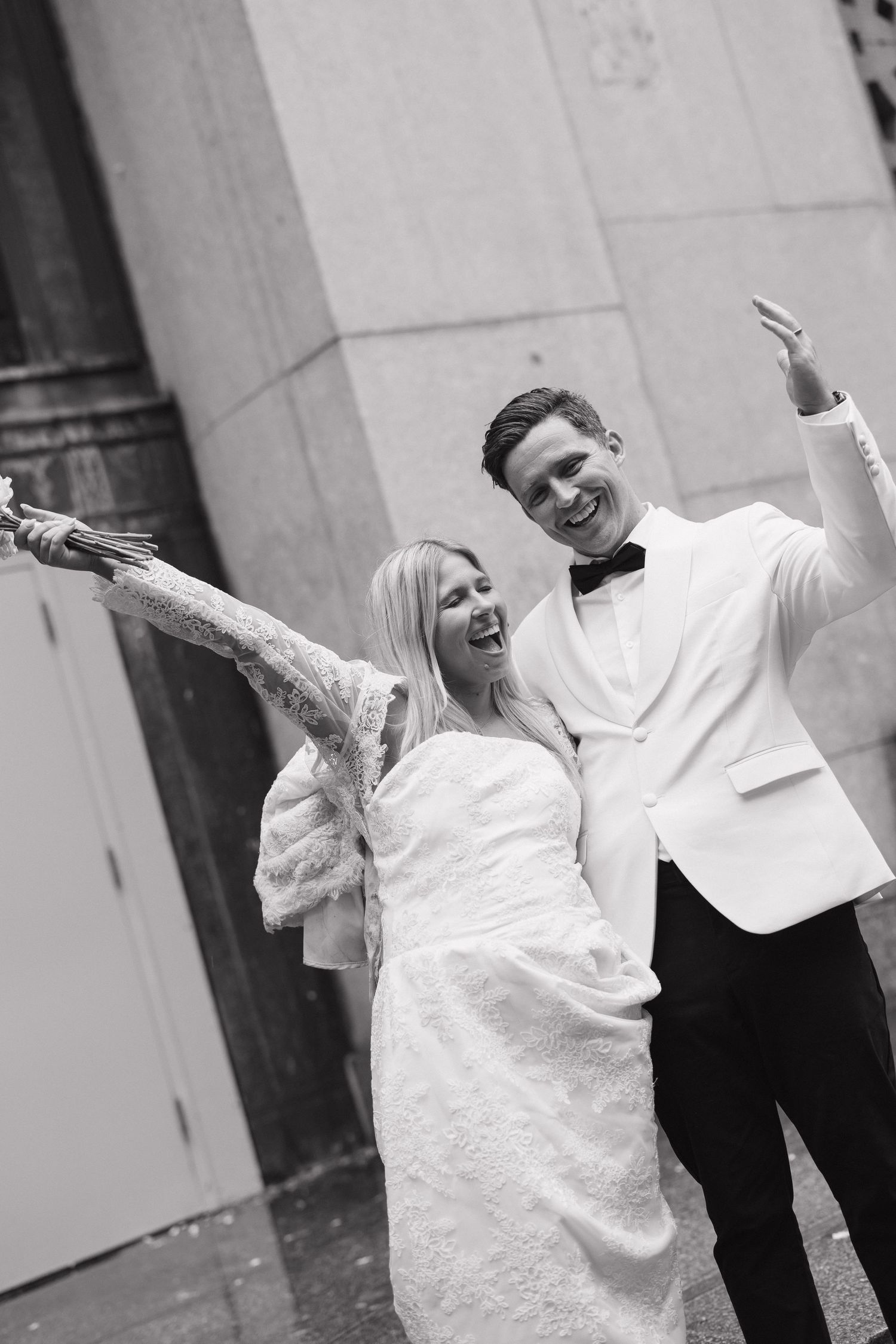 Two people in white outfits dancing playfully against a stone wall.
