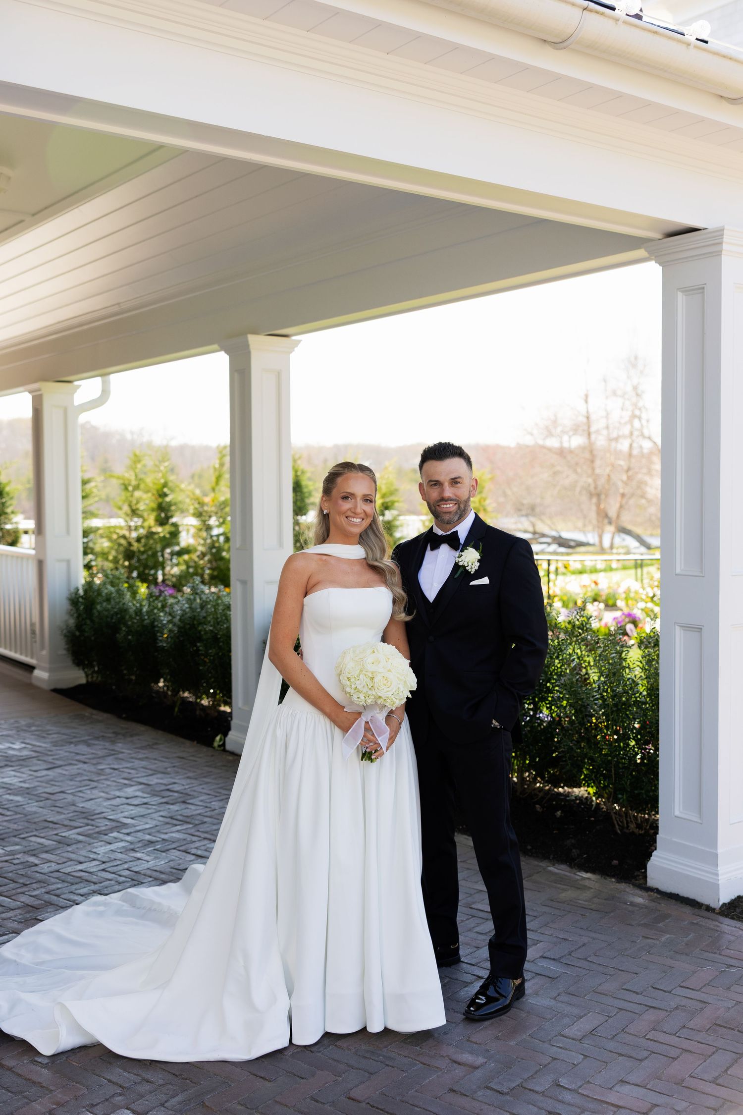 Bride in strapless white gown holding bouquet stands with groom in black tuxedo under elegant venue portico.