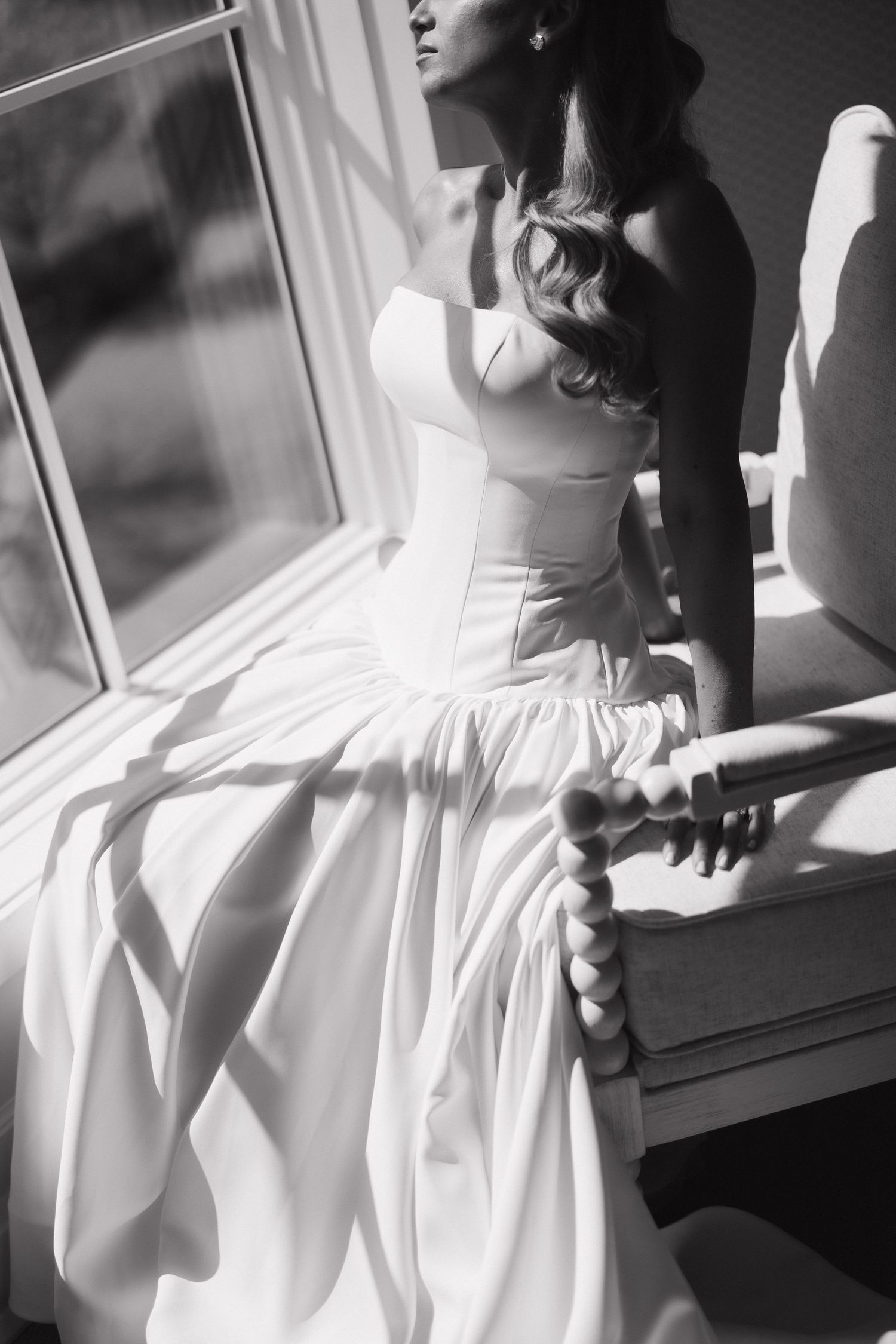 Black and white photo of a bride in a strapless ball gown sitting by a sunlit window.