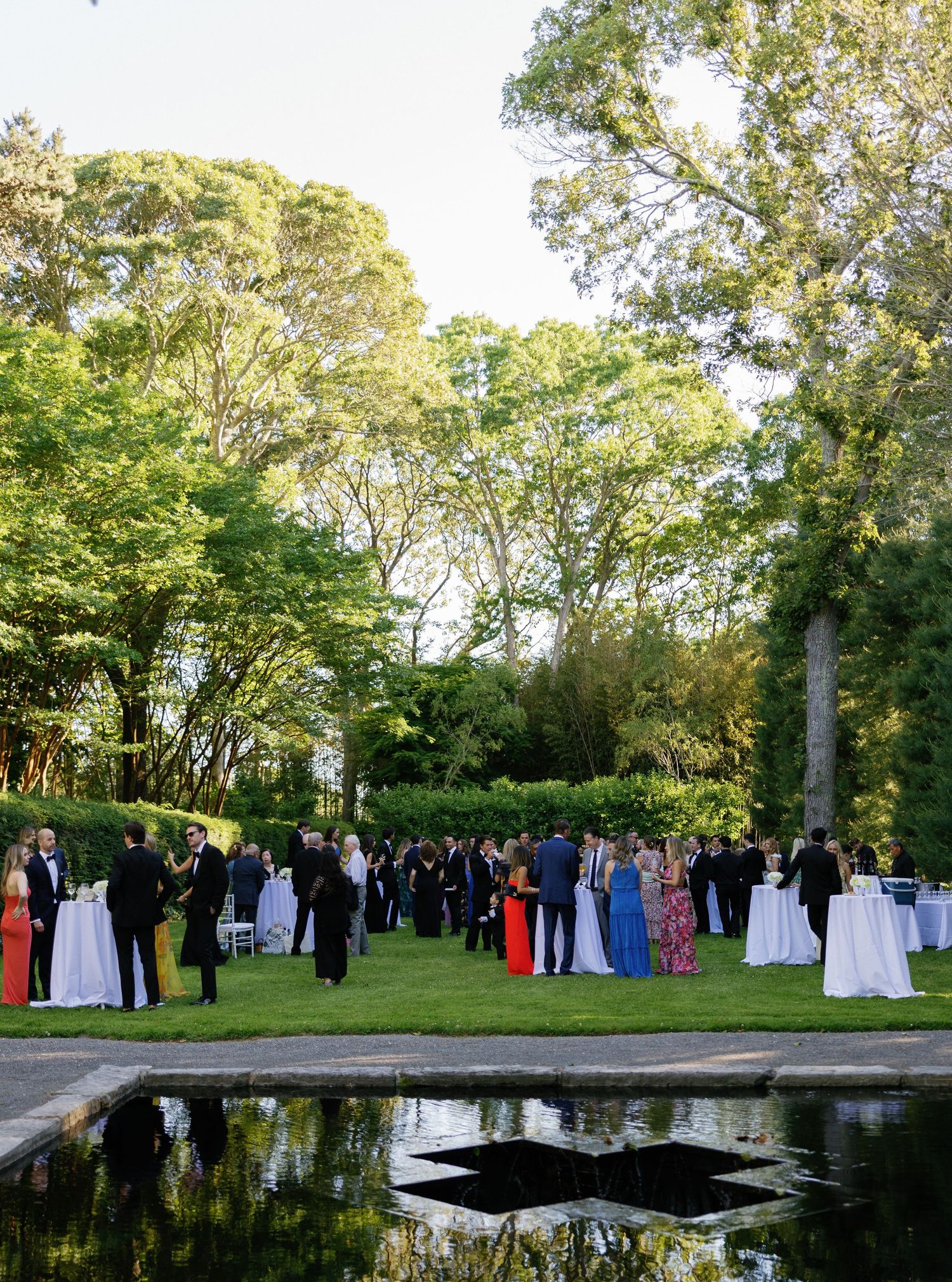 Wedding guests gather around a reflective pool during an outdoor garden ceremony.
