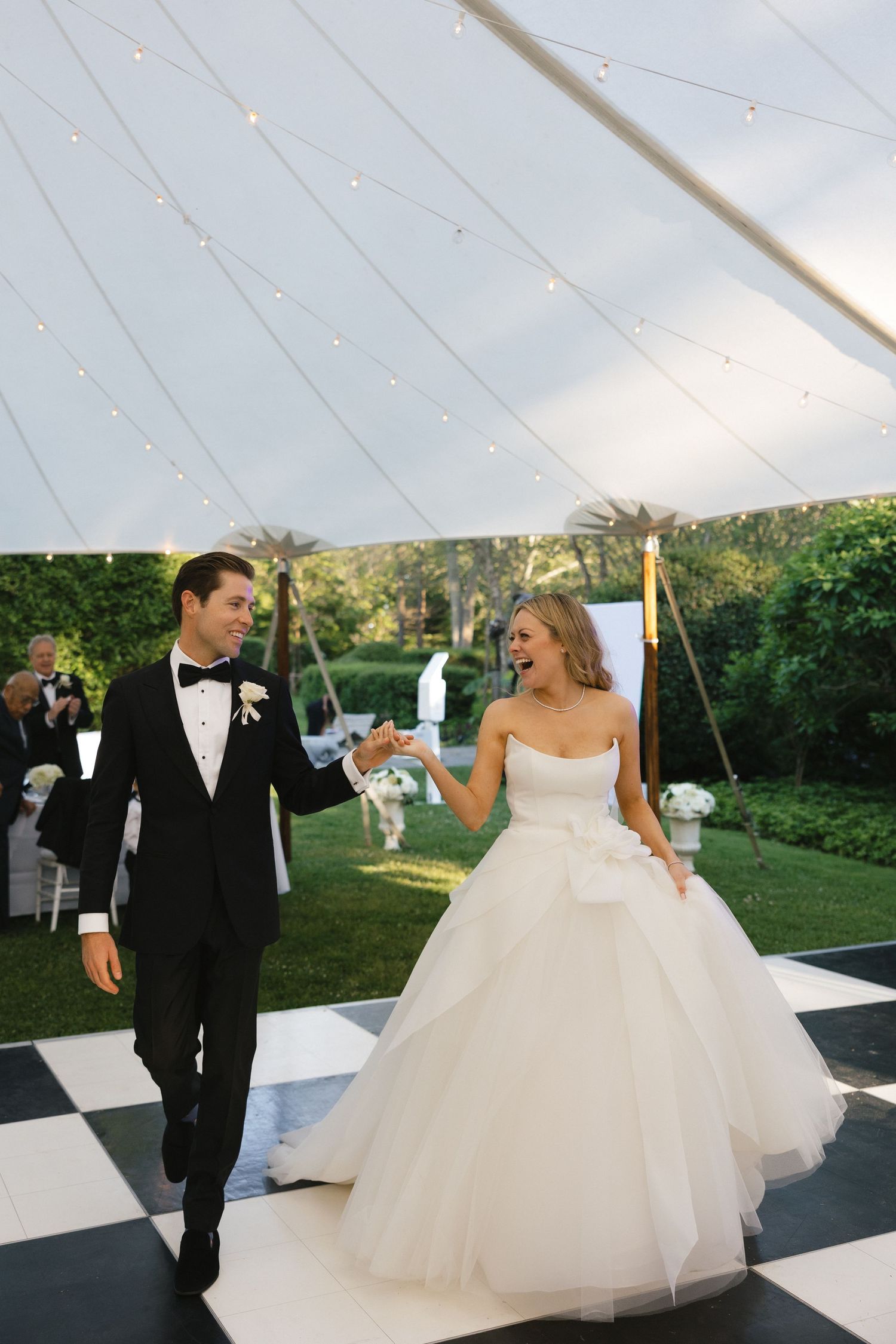 A couple shares their first dance on a black and white checkered floor under a tent with string lights.