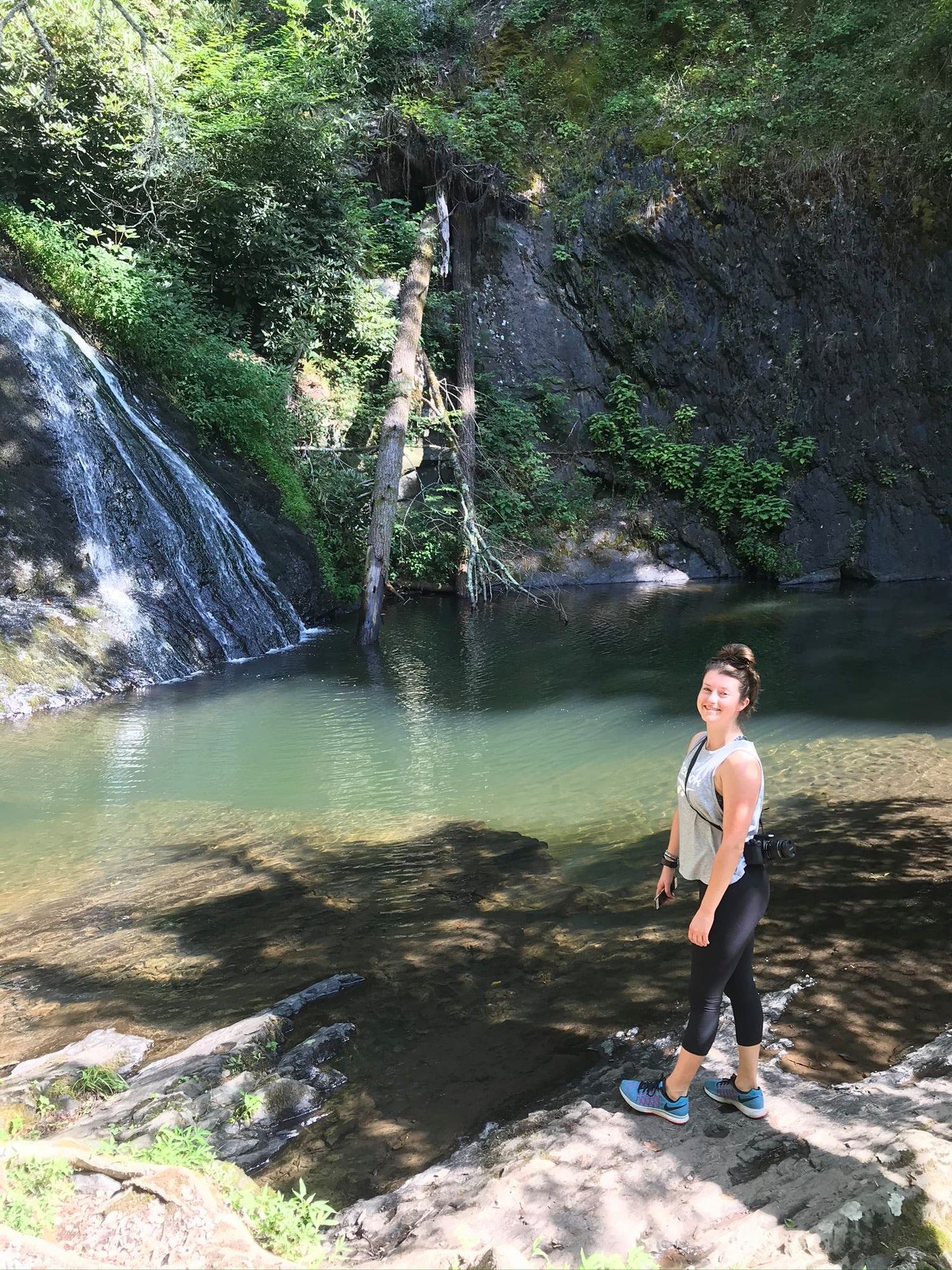 A person stands near a scenic waterfall in a lush forest setting.