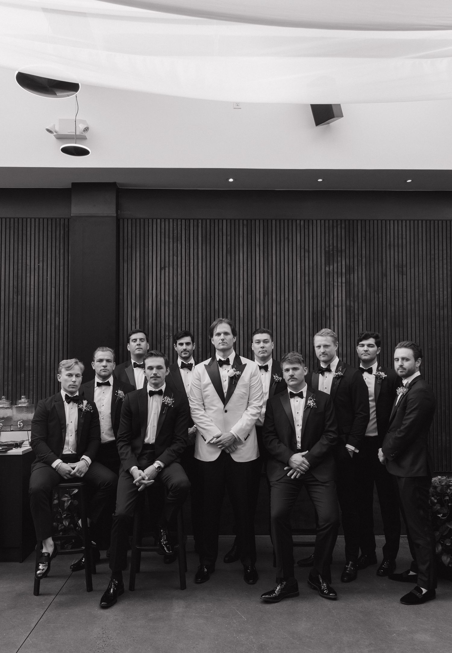 Black and white portrait of groomsmen in tuxedos posed formally against modern wall paneling.