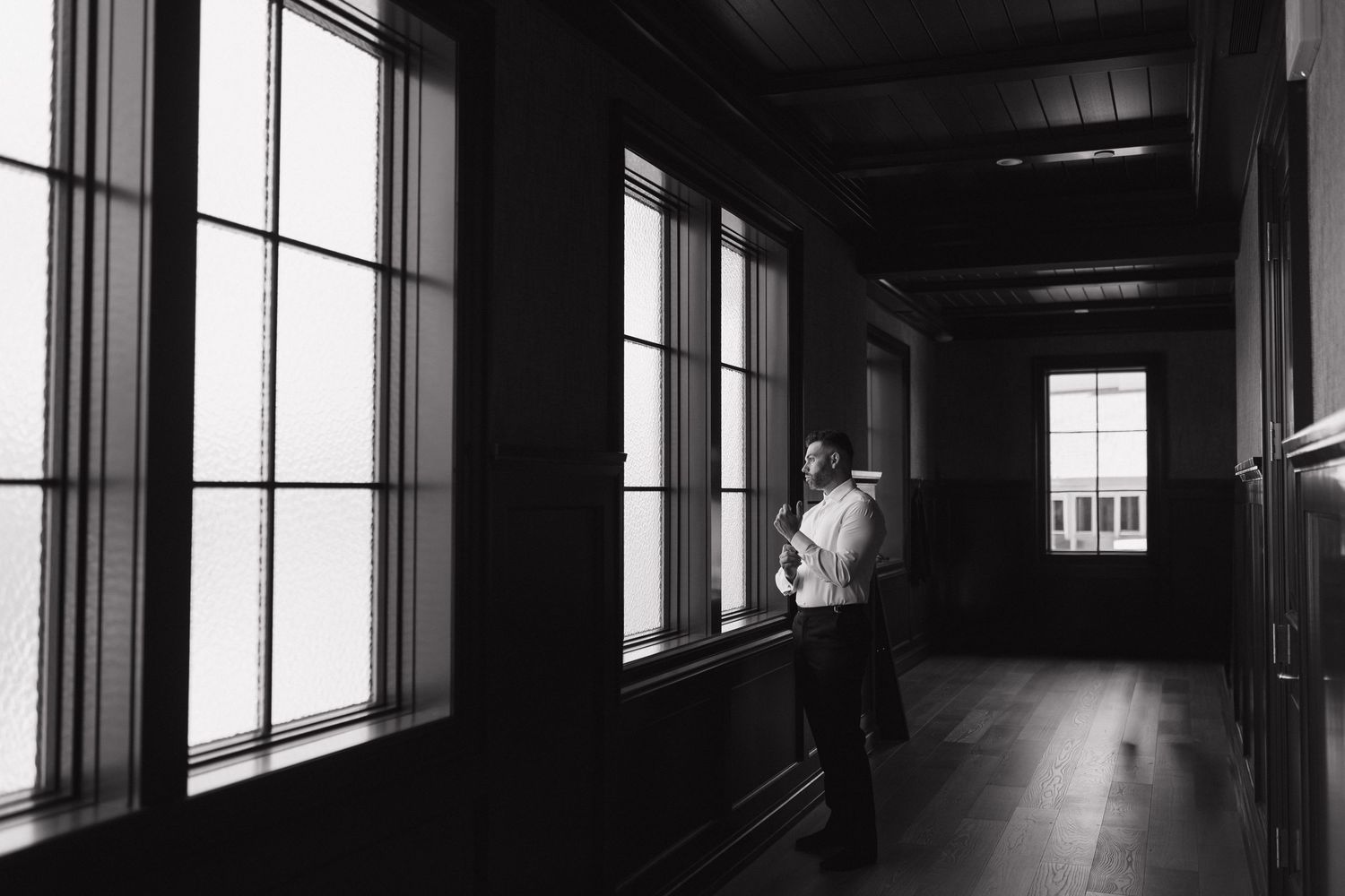 Black and white photo of a person standing in a dark hallway looking out large windows, creating a moody, contemplative atmosphere.