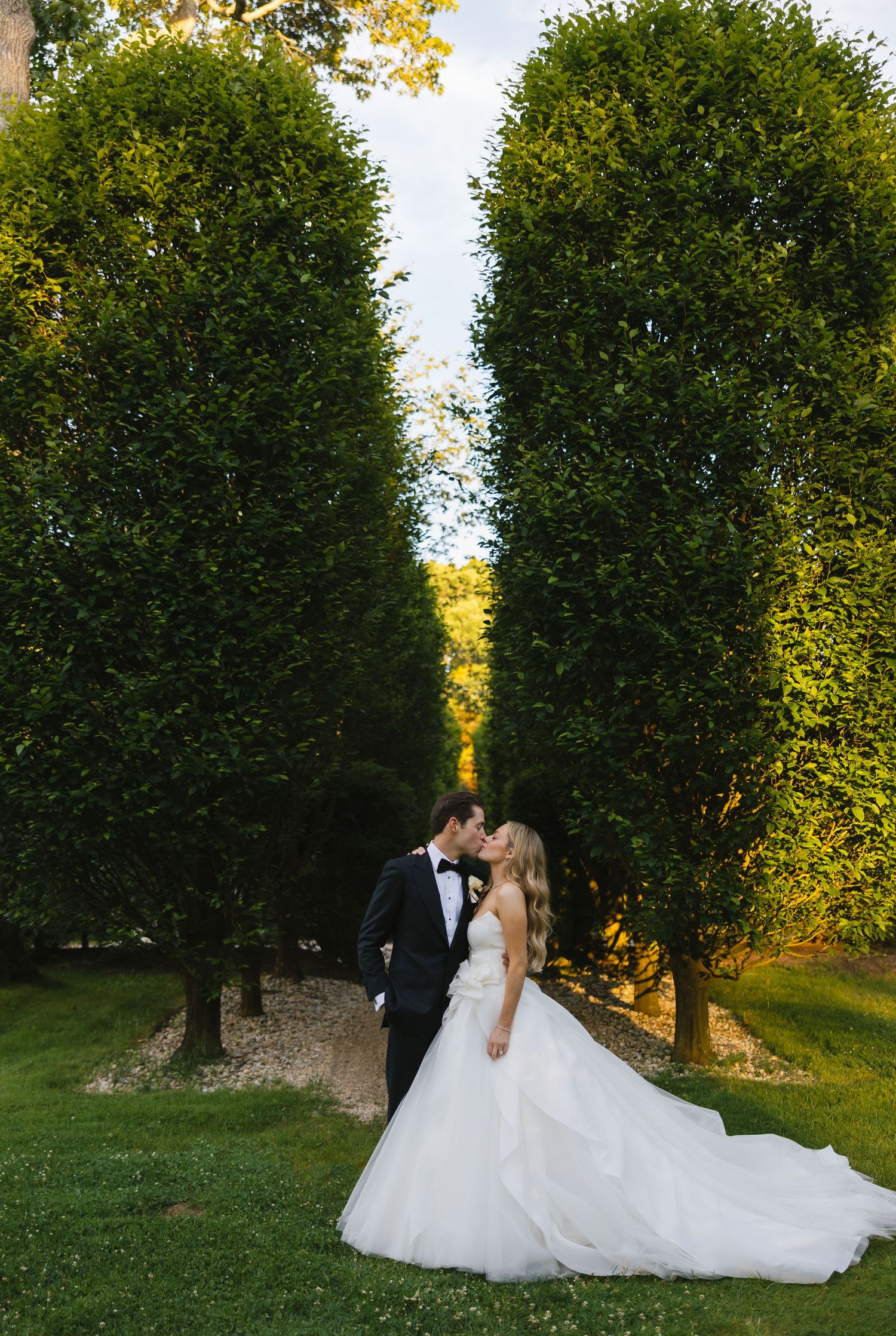 A couple in wedding attire poses for portraits among tall green trees at sunset.