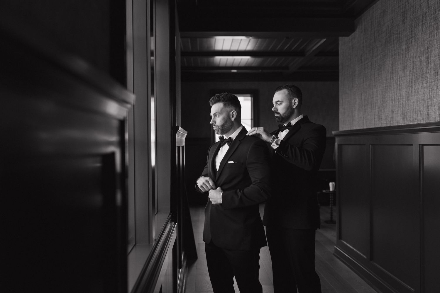 Two groomsmen in black tuxedos help each other get dressed in a dimly lit hallway before a wedding ceremony.