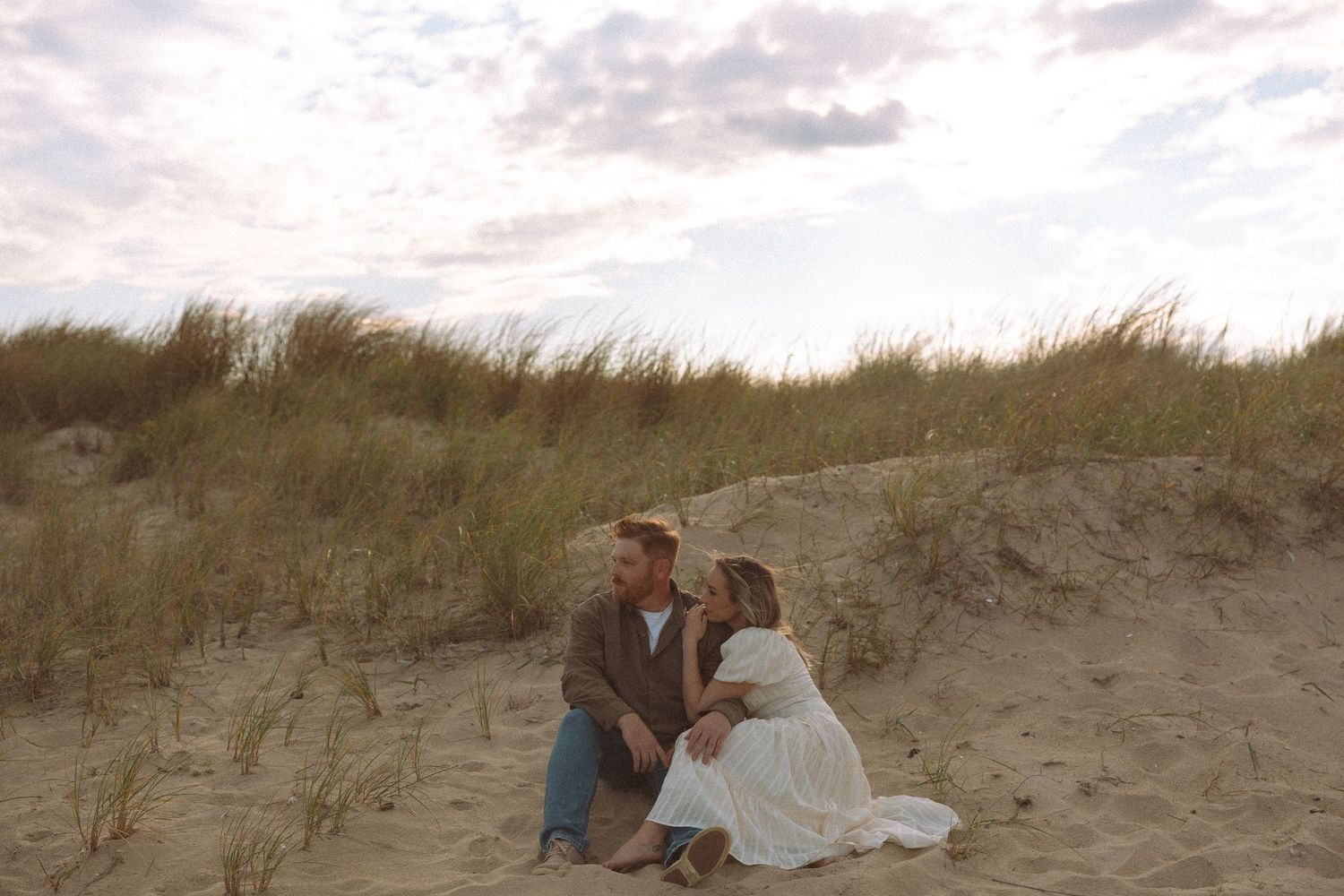 A couple shares a romantic kiss on a sandy beach dune during sunset, surrounded by coastal grass and soft lighting.