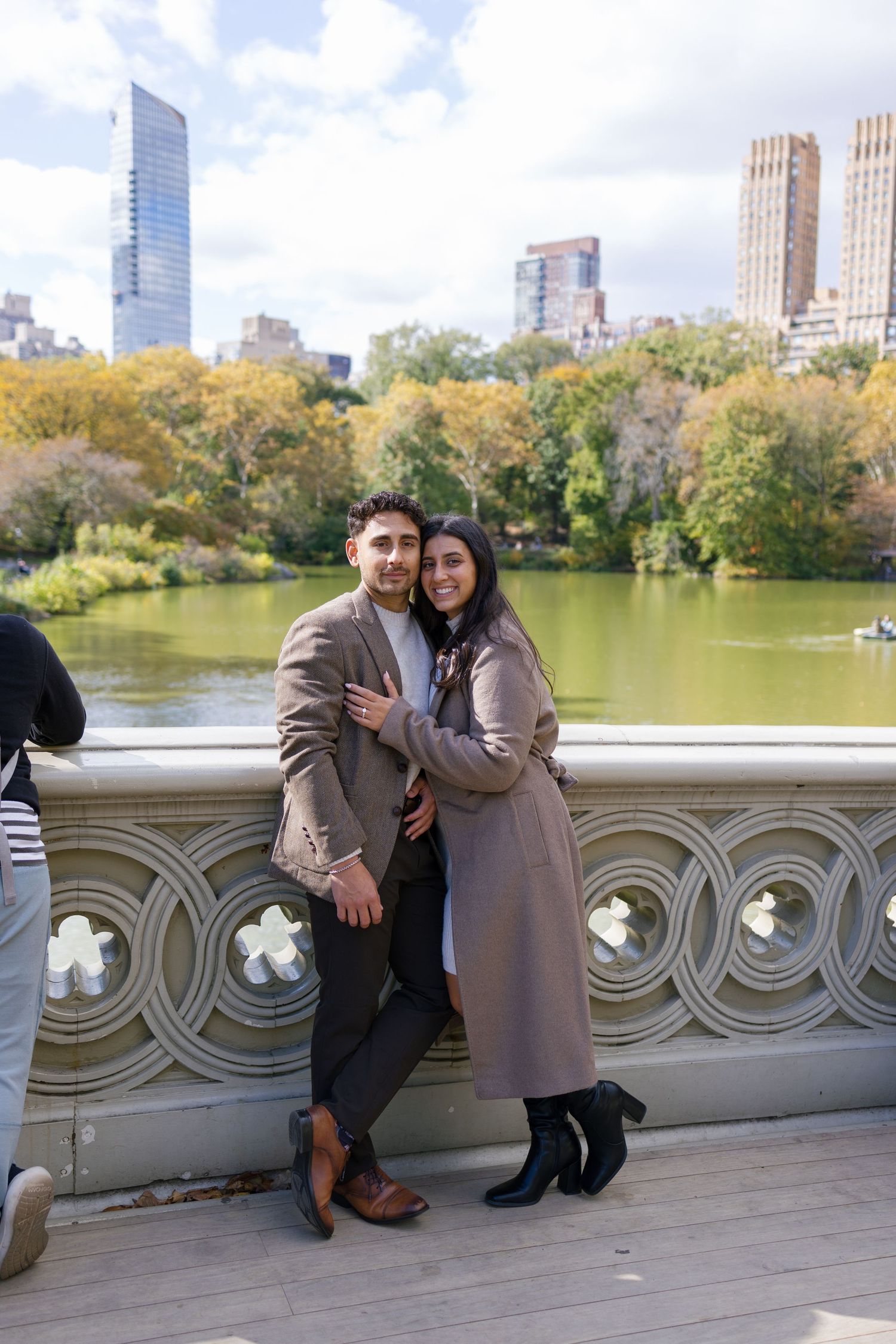 Couple sharing romantic moment on Bow Bridge with Central Park lake and city skyline in background.