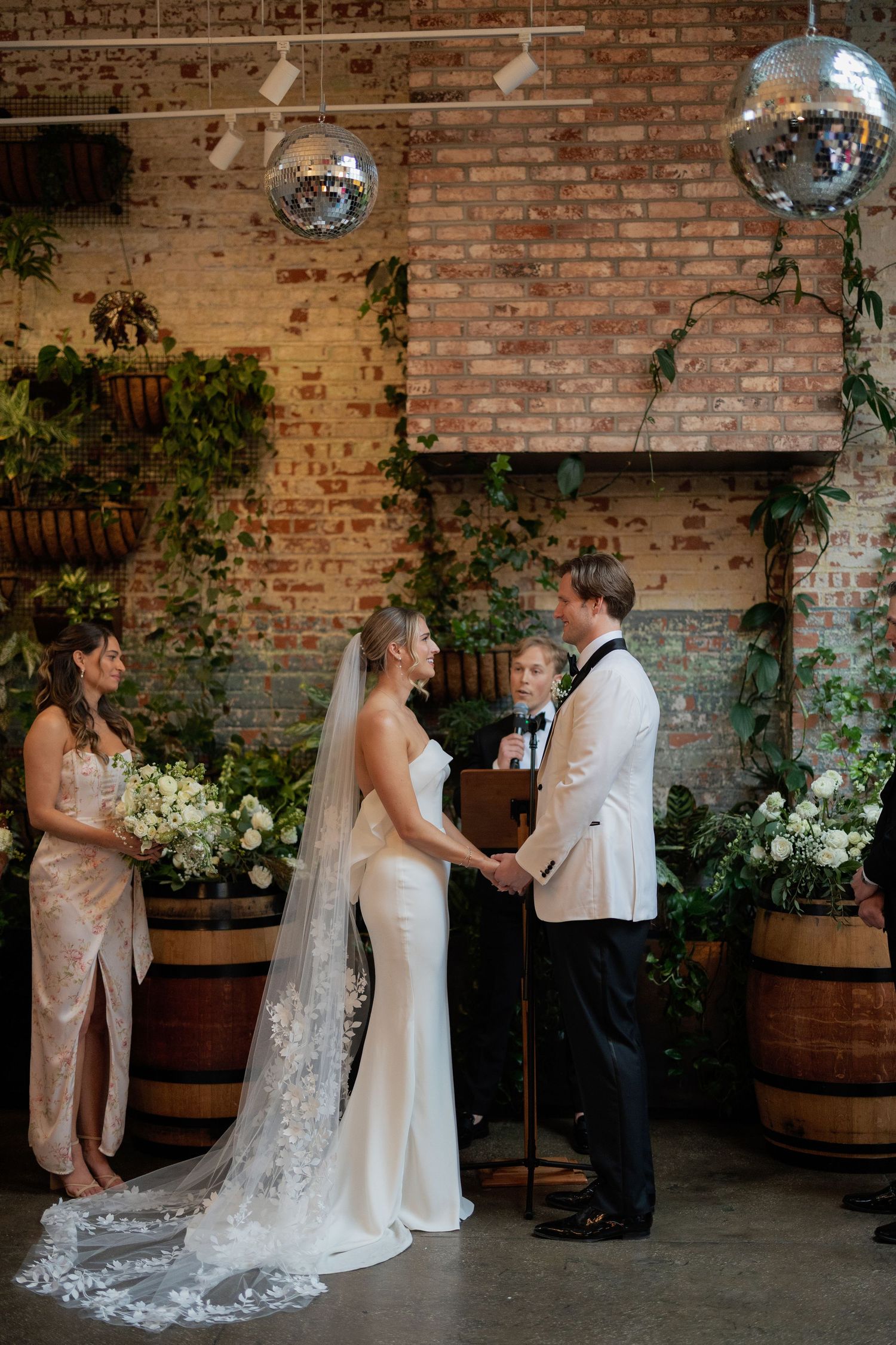 Wedding ceremony in a rustic brick venue with disco balls and wooden barrels decorated with greenery.
