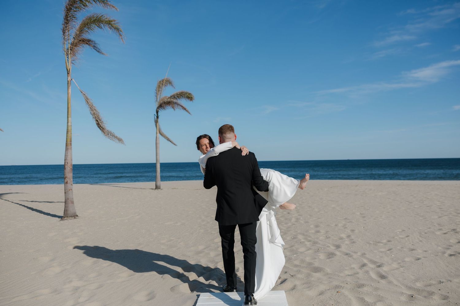 A couple embracing on a sunny beach with swaying palm trees and blue ocean backdrop.