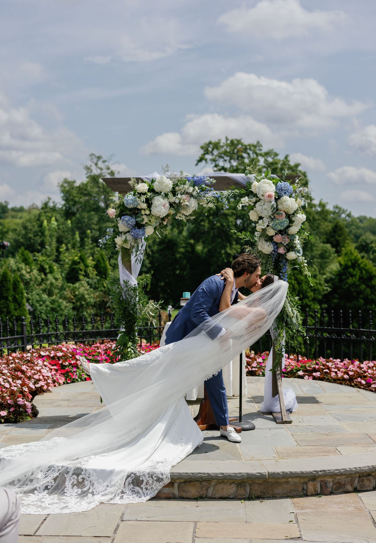 Wedding ceremony kiss under a floral arch with flowing veil in a garden setting.