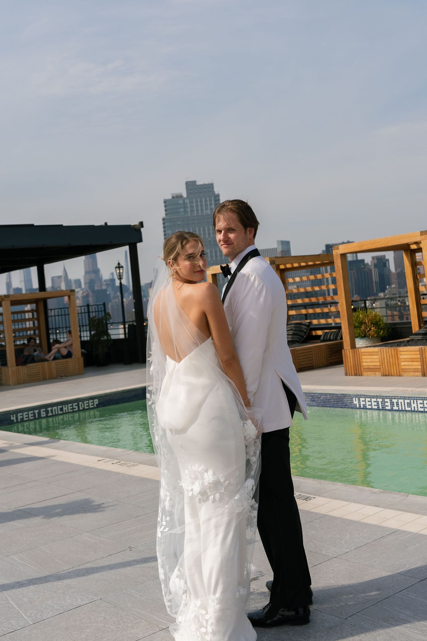 Newlyweds pose by a rooftop pool with city buildings visible in the background.