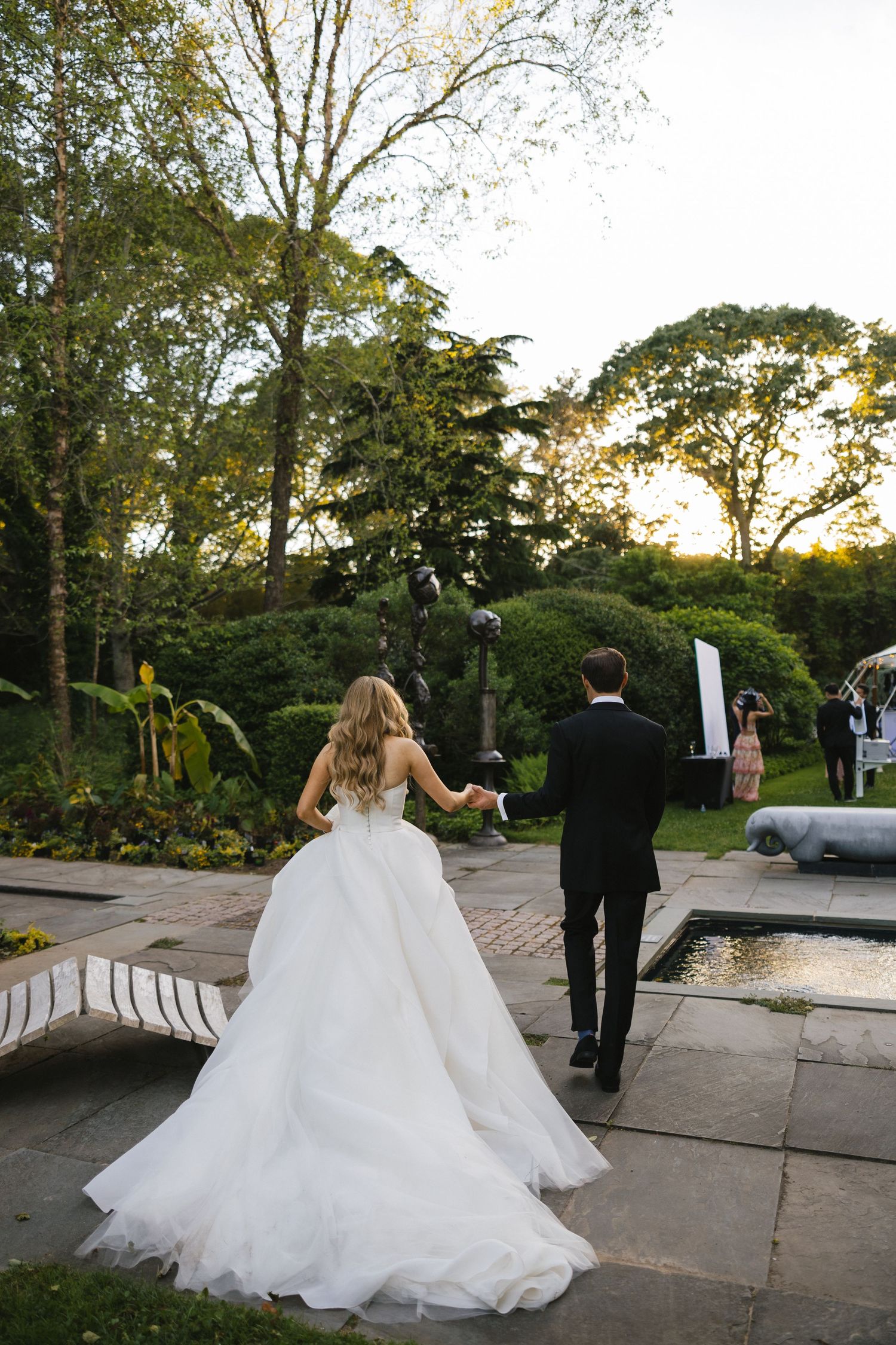 A couple in wedding attire walks through an elegant garden path at sunset.