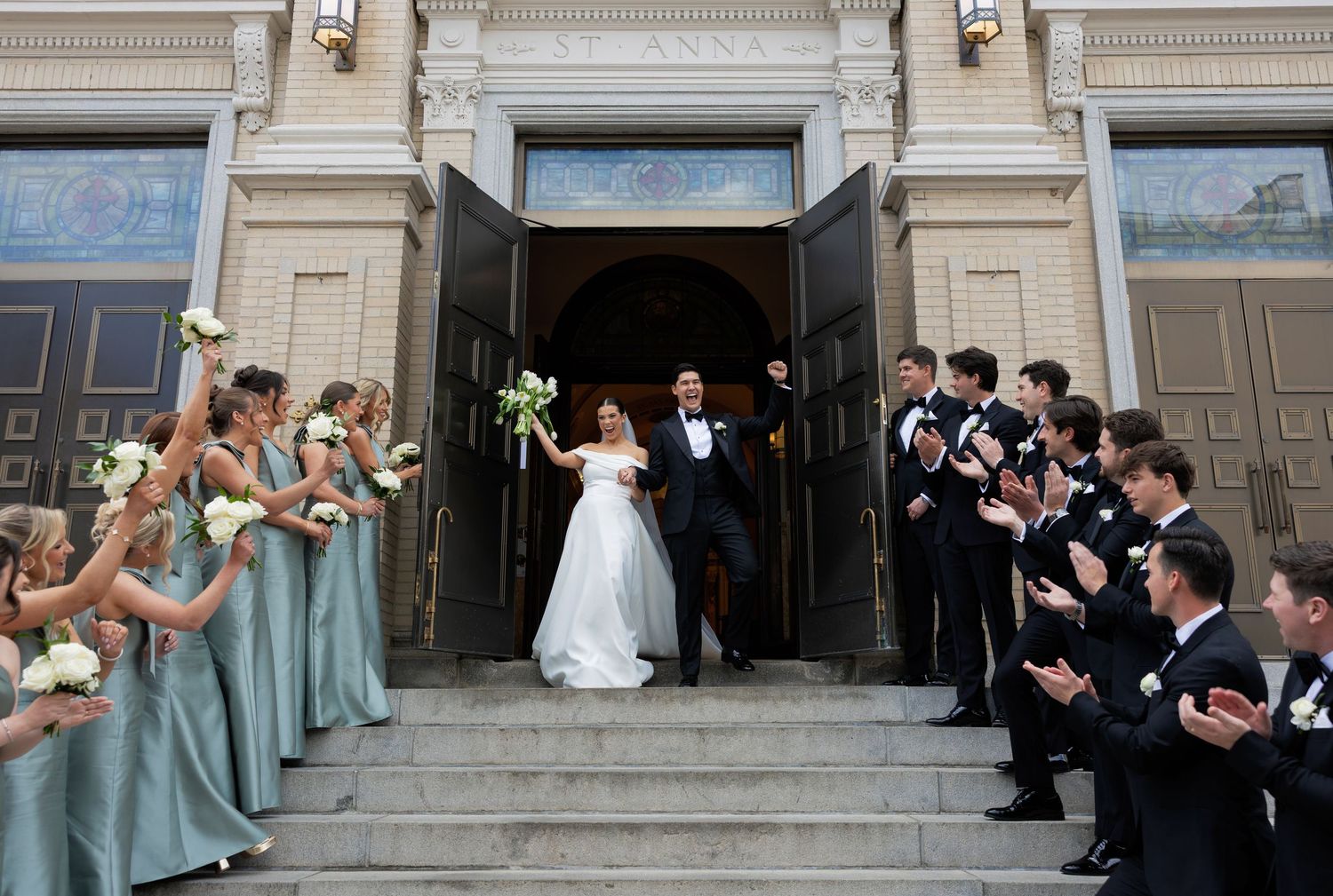 Bride and groom exit church doors as bridesmaids in sage green dresses and groomsmen in tuxedos cheer on the steps.