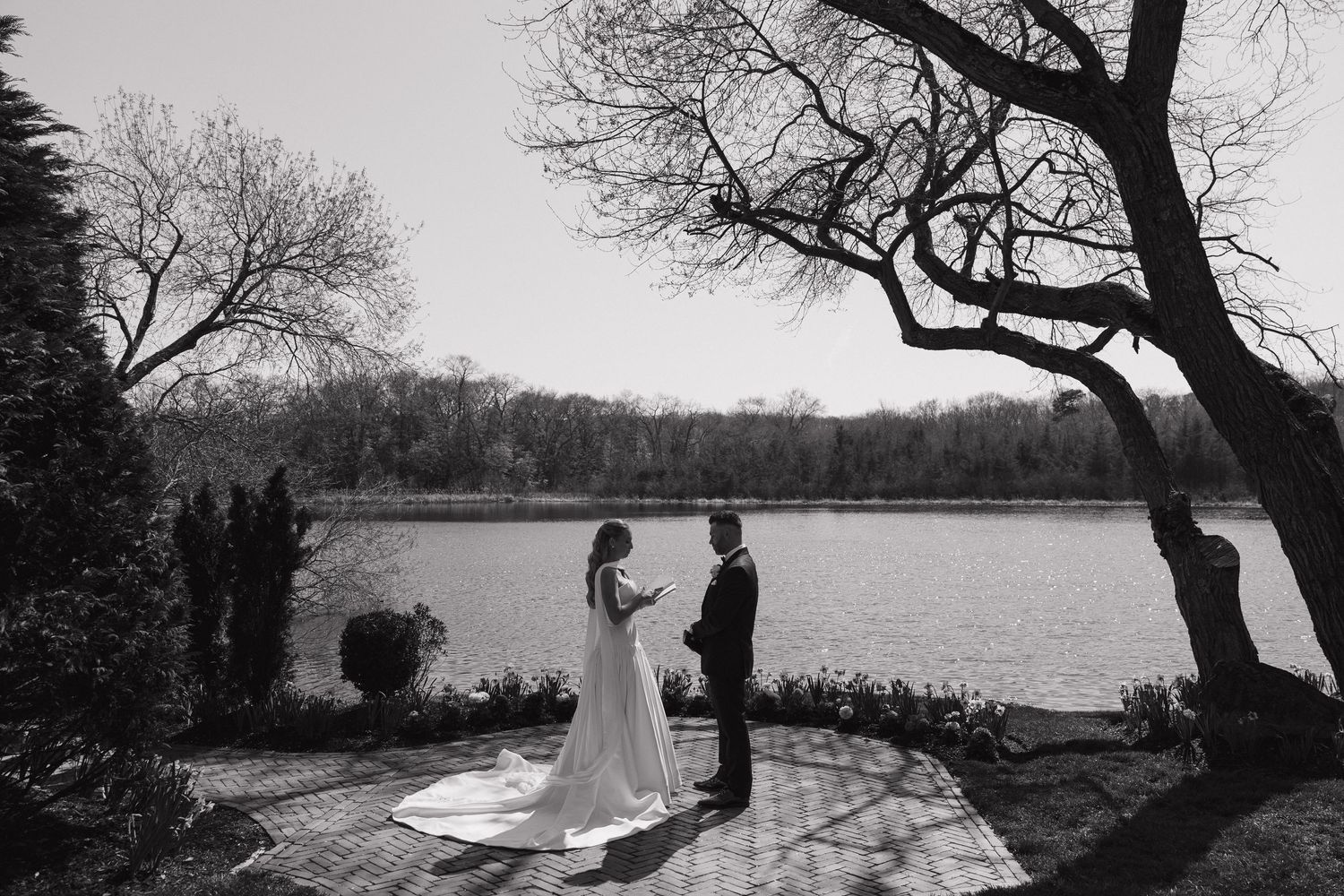 Black and white photo of bride and groom sharing first look by serene lakeside under bare trees.